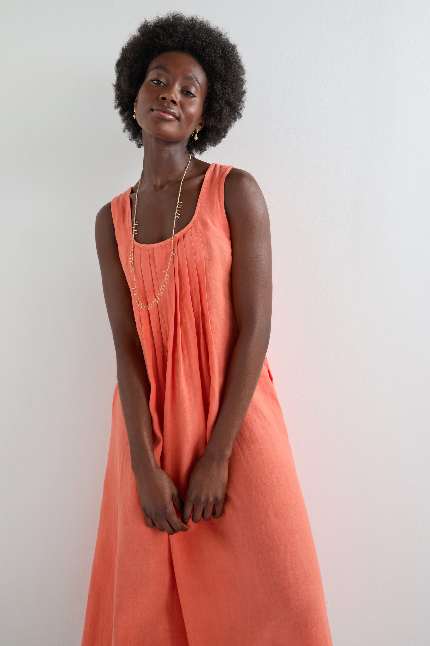 A woman stands against a plain white wall, wearing the Linen Pleat Front Sleeveless Dress in coral and a long necklace.