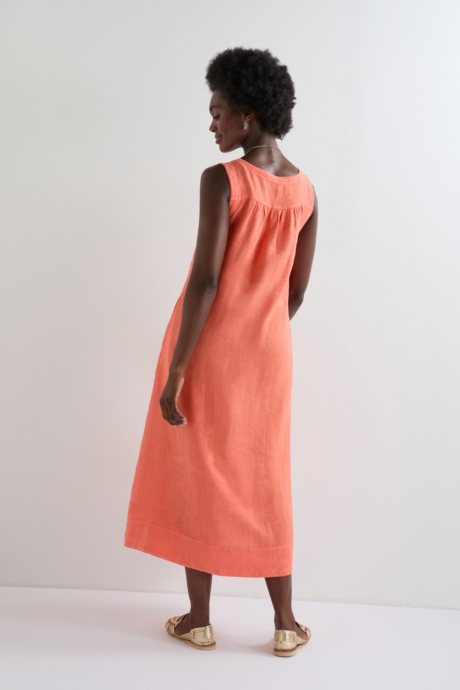 A woman stands facing away, modeling the Linen Pleat Front Sleeveless Dress in coral with gold sandals against a plain white background.