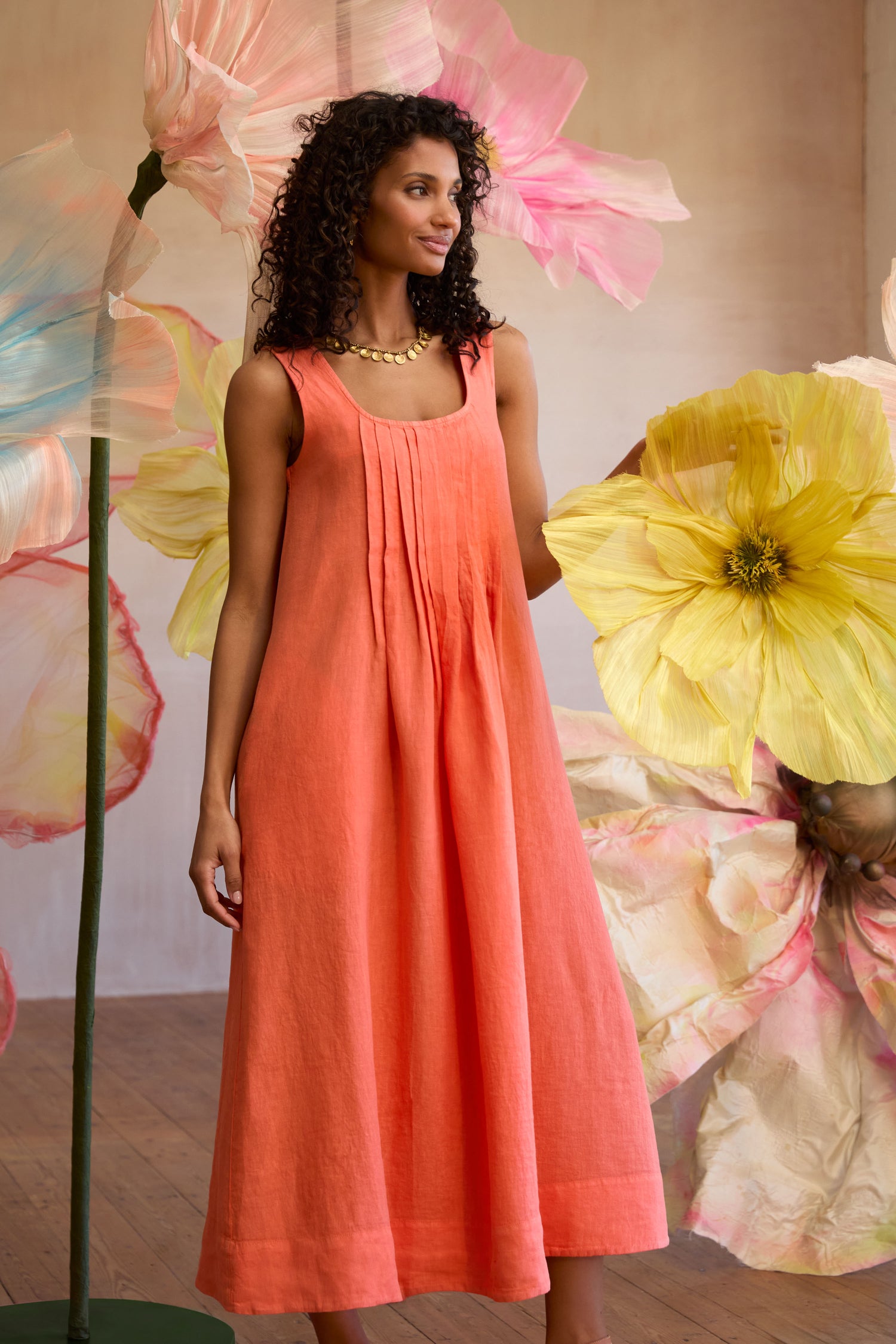 A woman wearing the Garment Dyed Linen Pleat Front Sleeveless Dress stands amid oversized pastel flowers on wooden floors, highlighting the comfort and style of breathable linen.
