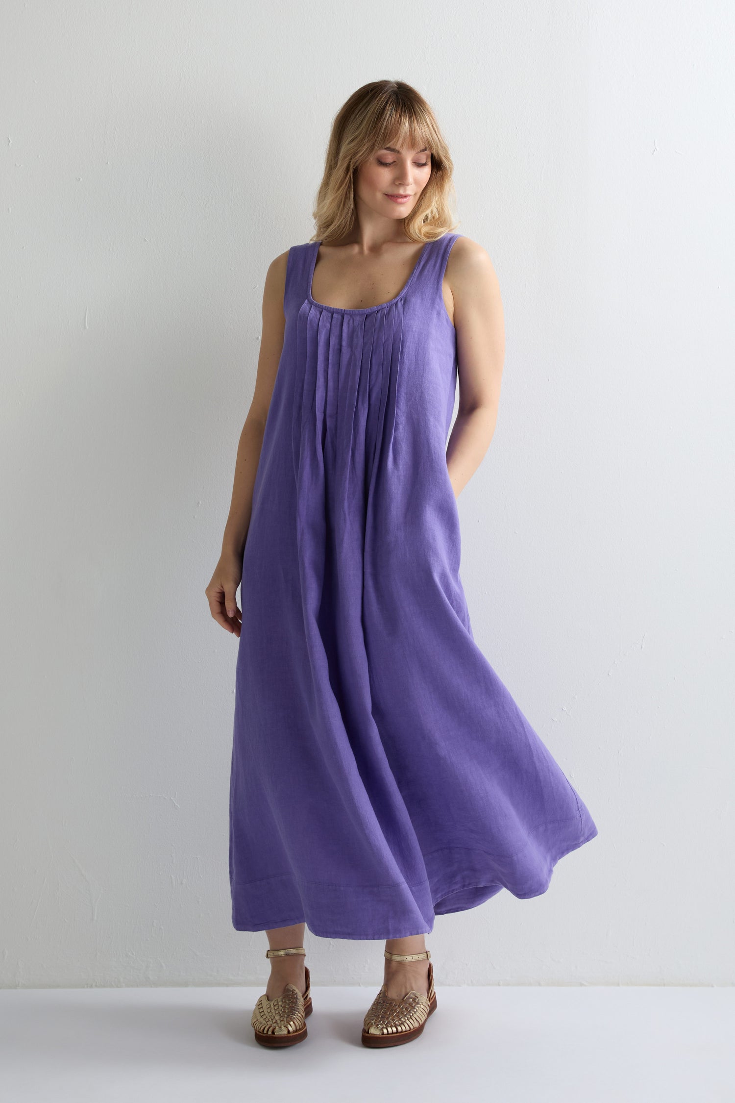 A woman stands against a plain white background wearing a Linen Pleat Front Sleeveless Dress in purple and tan woven sandals, her hands in the dress's pockets.