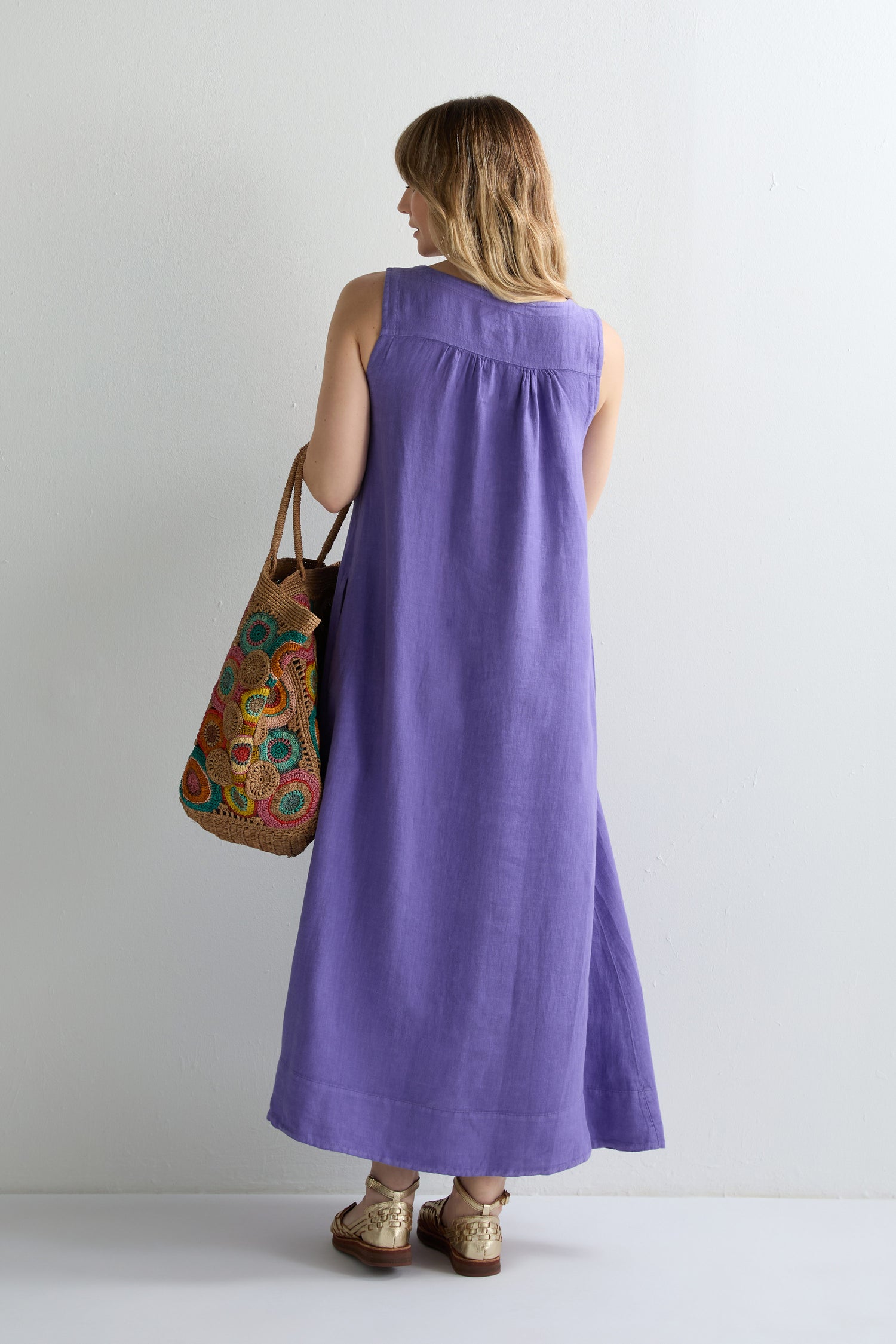 A person stands facing away, wearing a Linen Pleat Front Sleeveless Dress in purple and gold sandals, holding a brown woven bag with colorful circular patterns.