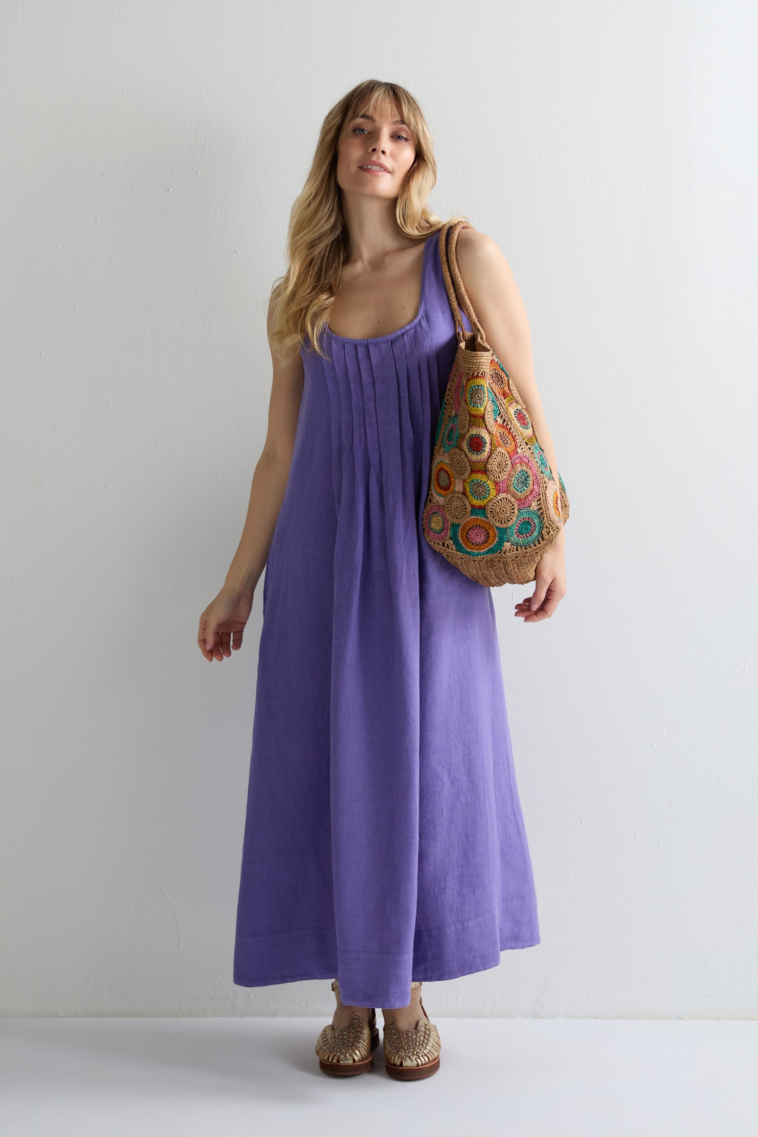 A woman stands against a plain wall wearing the Linen Pleat Front Sleeveless Dress in purple, brown woven shoes, and carrying a large multicolored crochet bag on her shoulder.