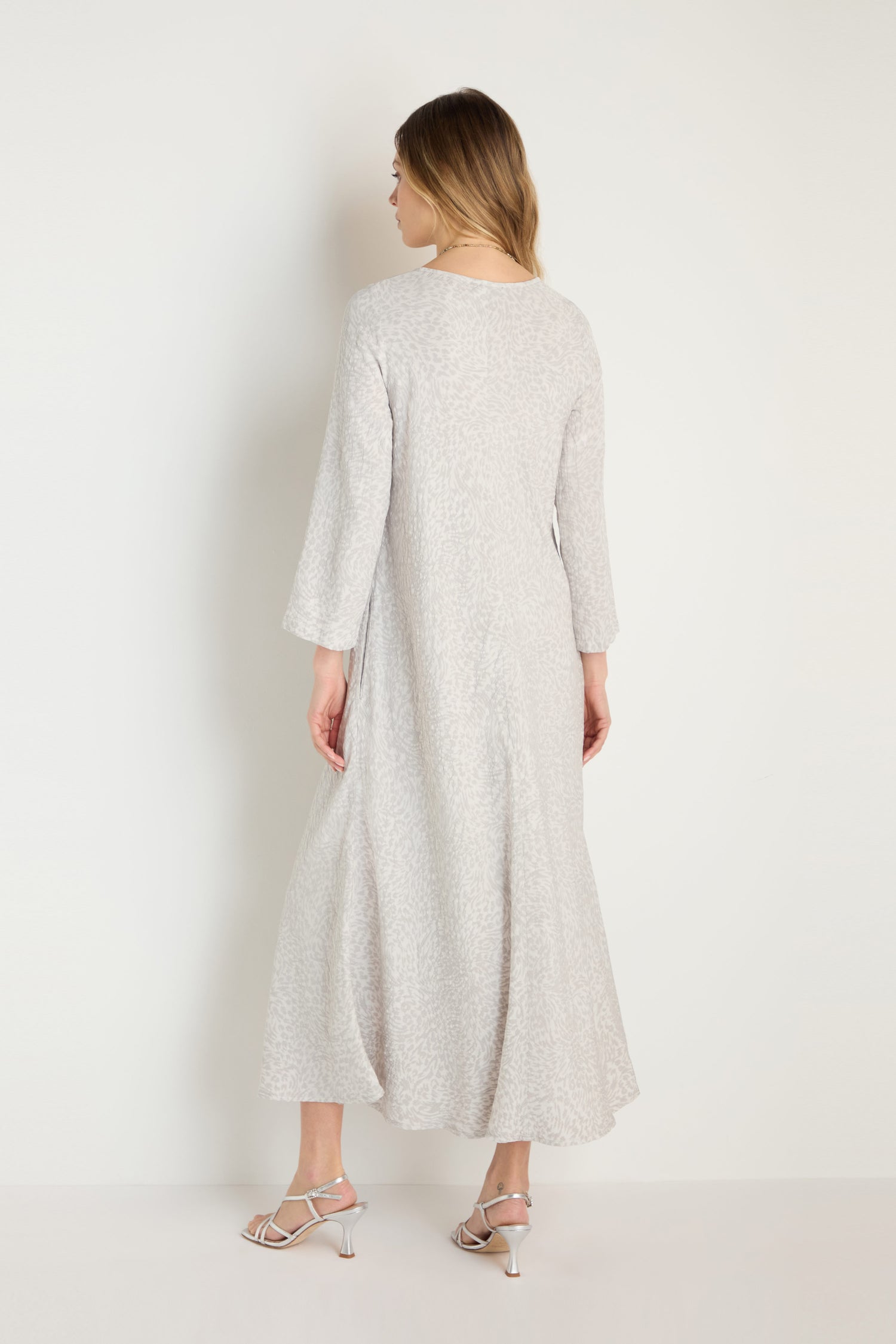 A woman faces away, wearing the Animal Textured Jacquard Maxi Dress in light gray with a subtle pattern and white heeled sandals, set against a plain light background.