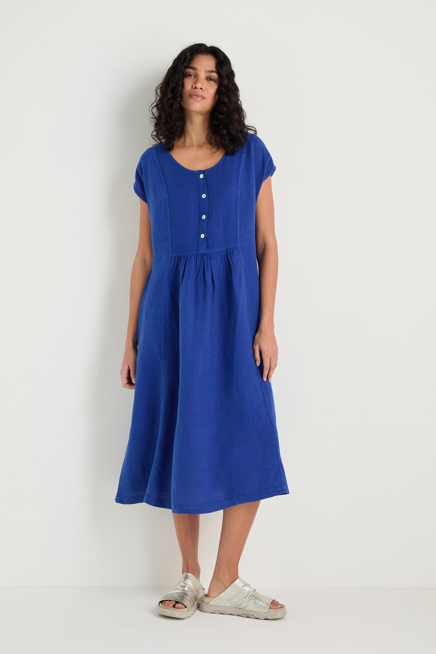 Wearing the Linen Button Cerulean Placket Dress with short sleeves and a fit-and-flare silhouette, paired with beige sandals, a person stands against a plain white background.