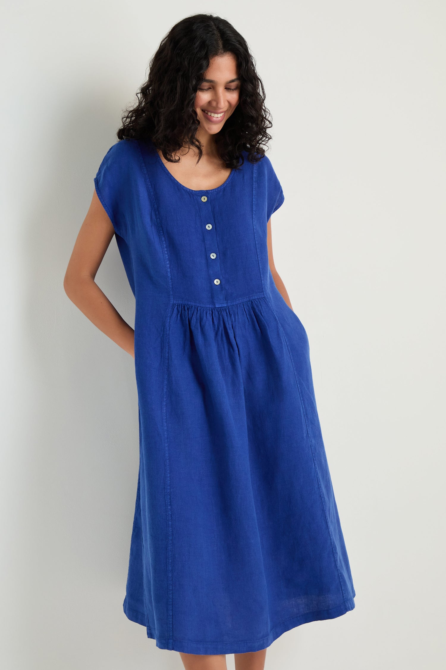 A woman stands by a light wall wearing the Linen Button Cerulean Placket Dress—a knee-length, short-sleeve, fit-and-flare blue dress with a front button placket and her hands in the pockets.