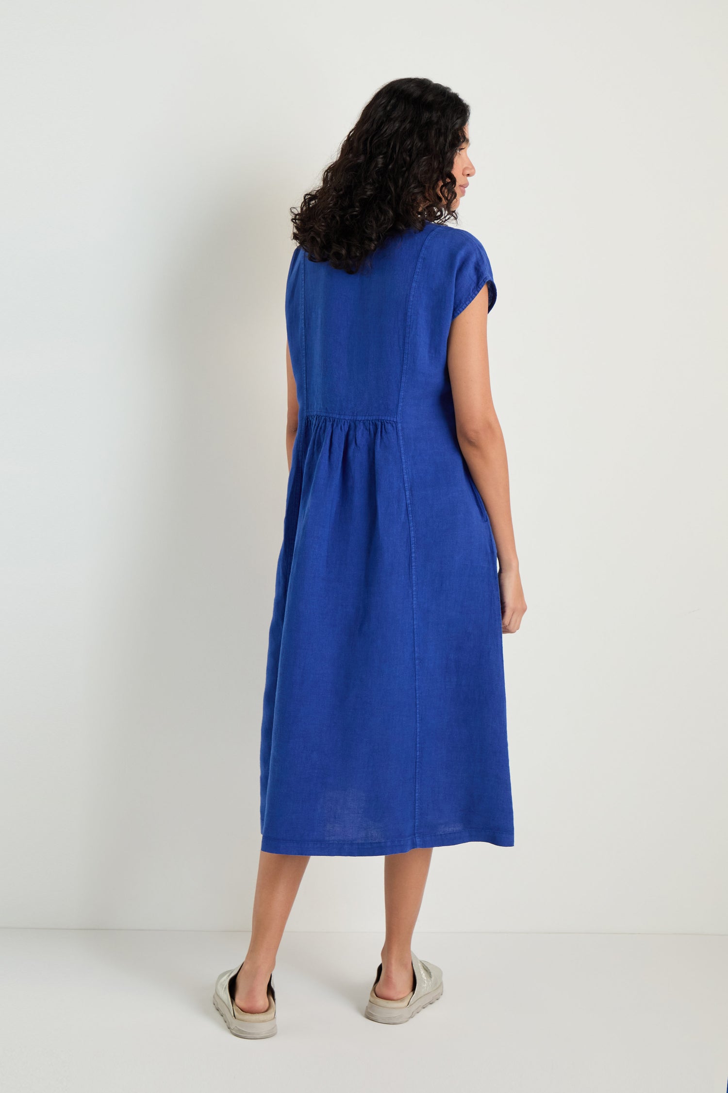 A woman with curly hair, seen from behind, wears the Linen Button Cerulean Placket Dress and white slip-on shoes against a plain white background.