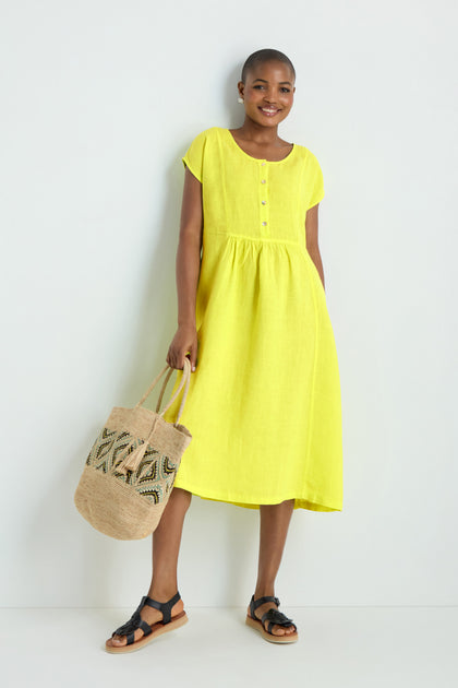 A person wearing the Linen Button Lemon Placket Dress, holding a woven tote bag and black sandals, stands against a plain white background.