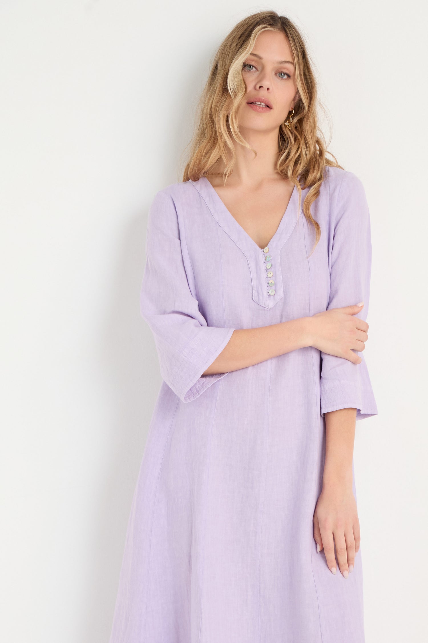 A woman with long blonde hair wears the Linen Seam Pansy Flare Dress—a light purple, V-neck, long-sleeve flare dress with panel seams and decorative buttons—standing against a plain white background.