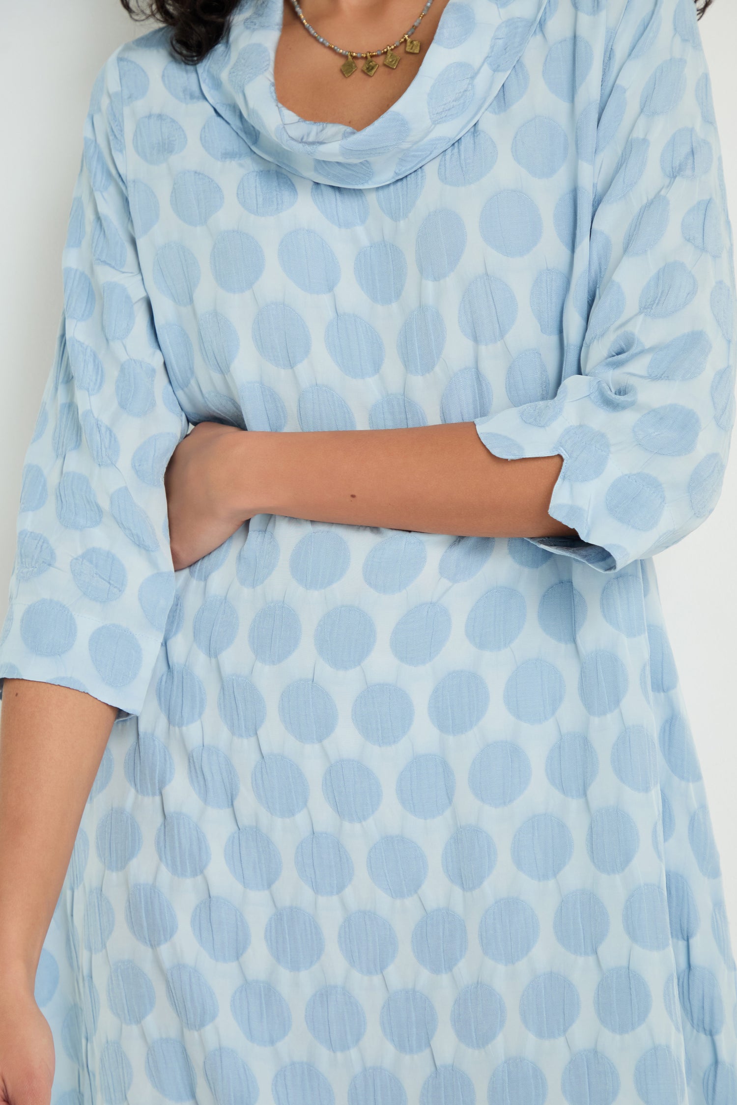 A person wears the Spot Textured Jacquard Bubble Dress, a light blue, long-sleeved dress with large polka dots, standing with one arm bent across their stomach.