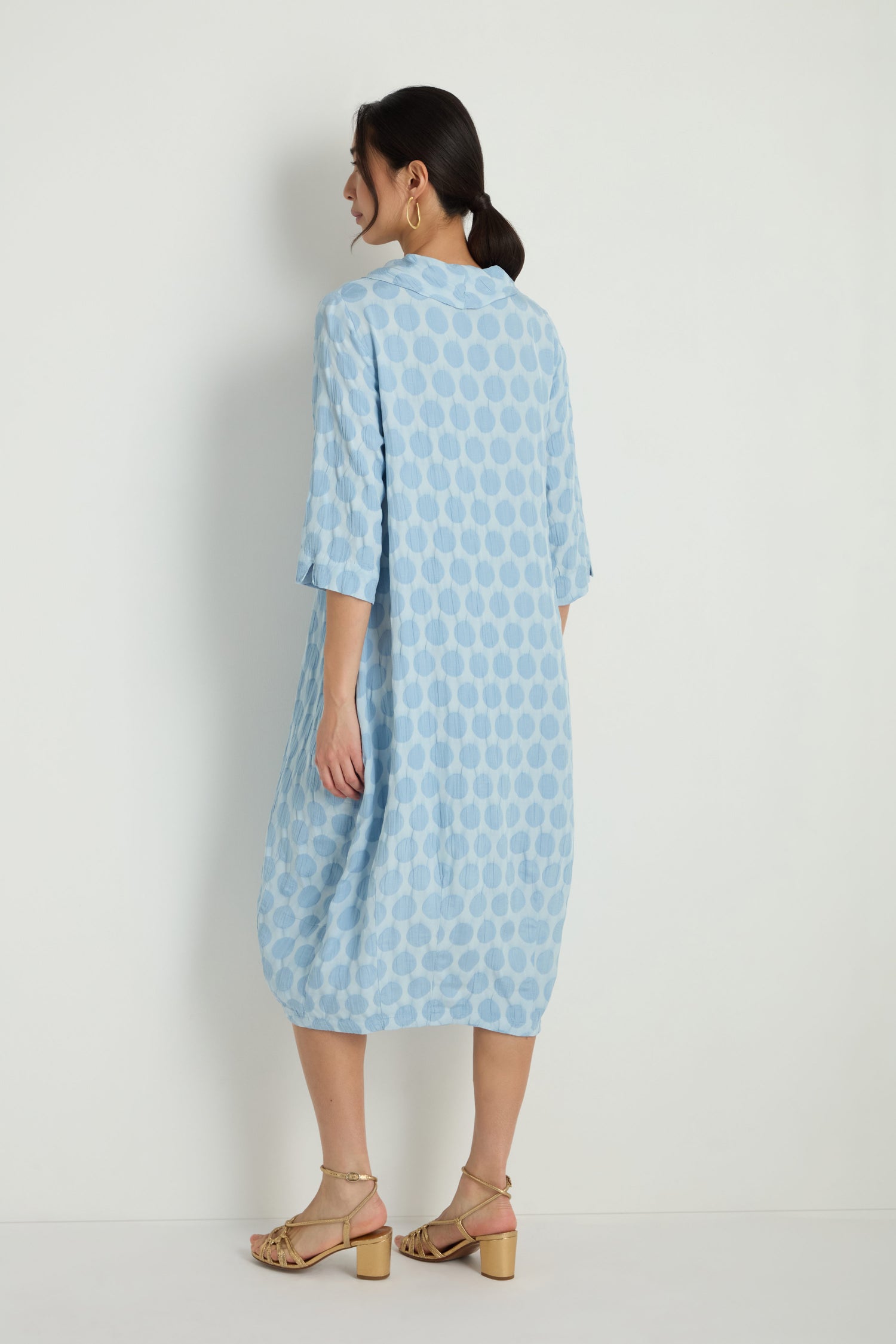 A woman smiles and holds the sides of her Spot Textured Jacquard Bubble Dress, a light blue, midi-length garment with a polka dot pattern, while standing against a plain white background.