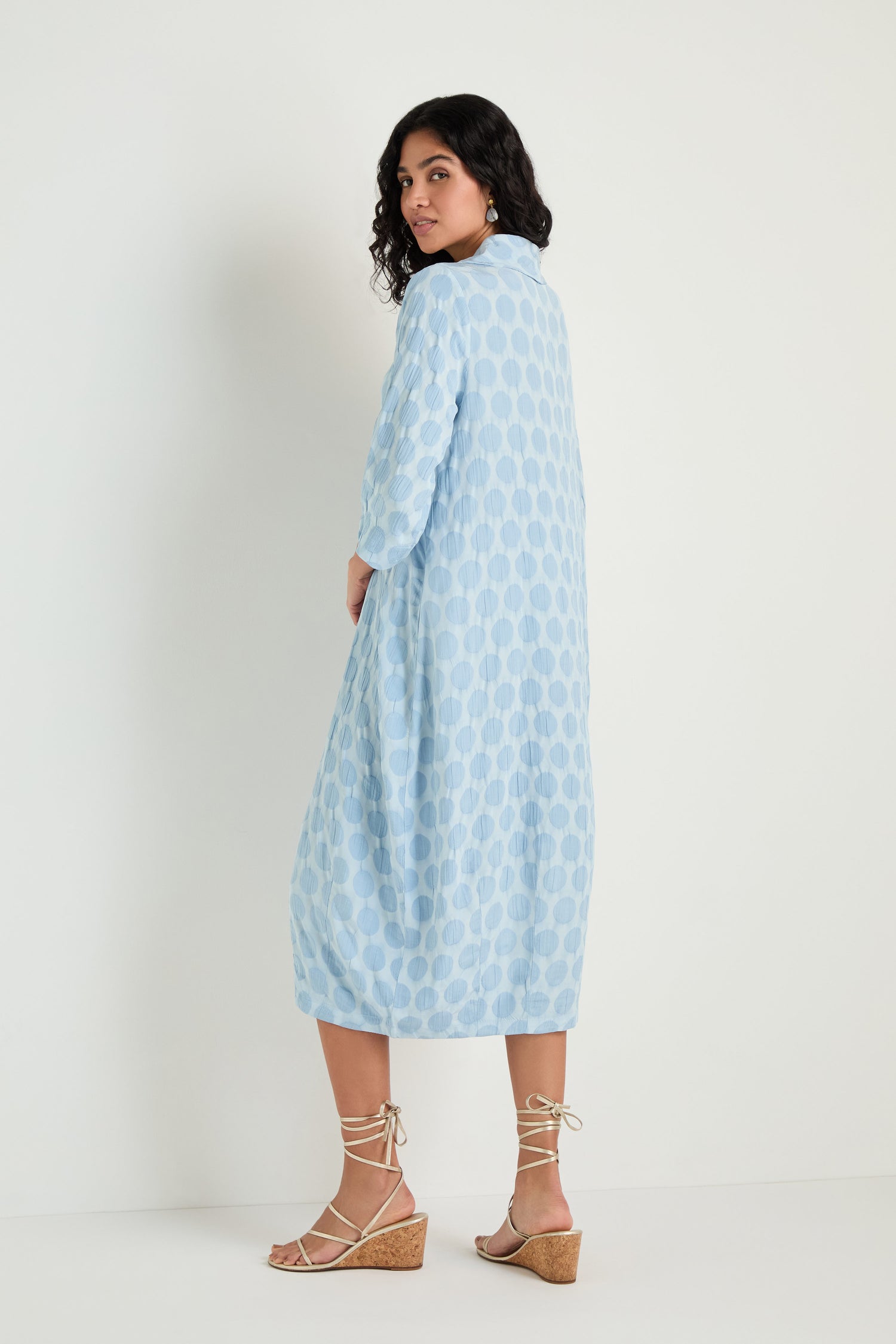 A woman stands against a white background wearing the Spot Textured Jacquard Bubble Dress in light blue with polka dots, styled with beige wedge sandals, looking over her shoulder.