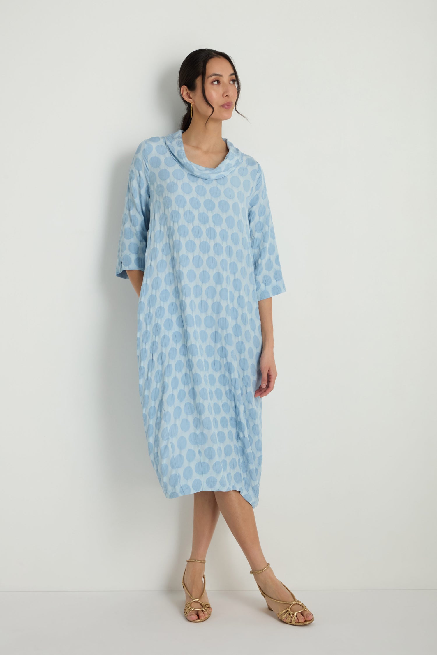 A woman stands against a white background wearing the Spot Textured Jacquard Bubble Dress in light blue with polka dots, styled with beige wedge sandals, looking over her shoulder.