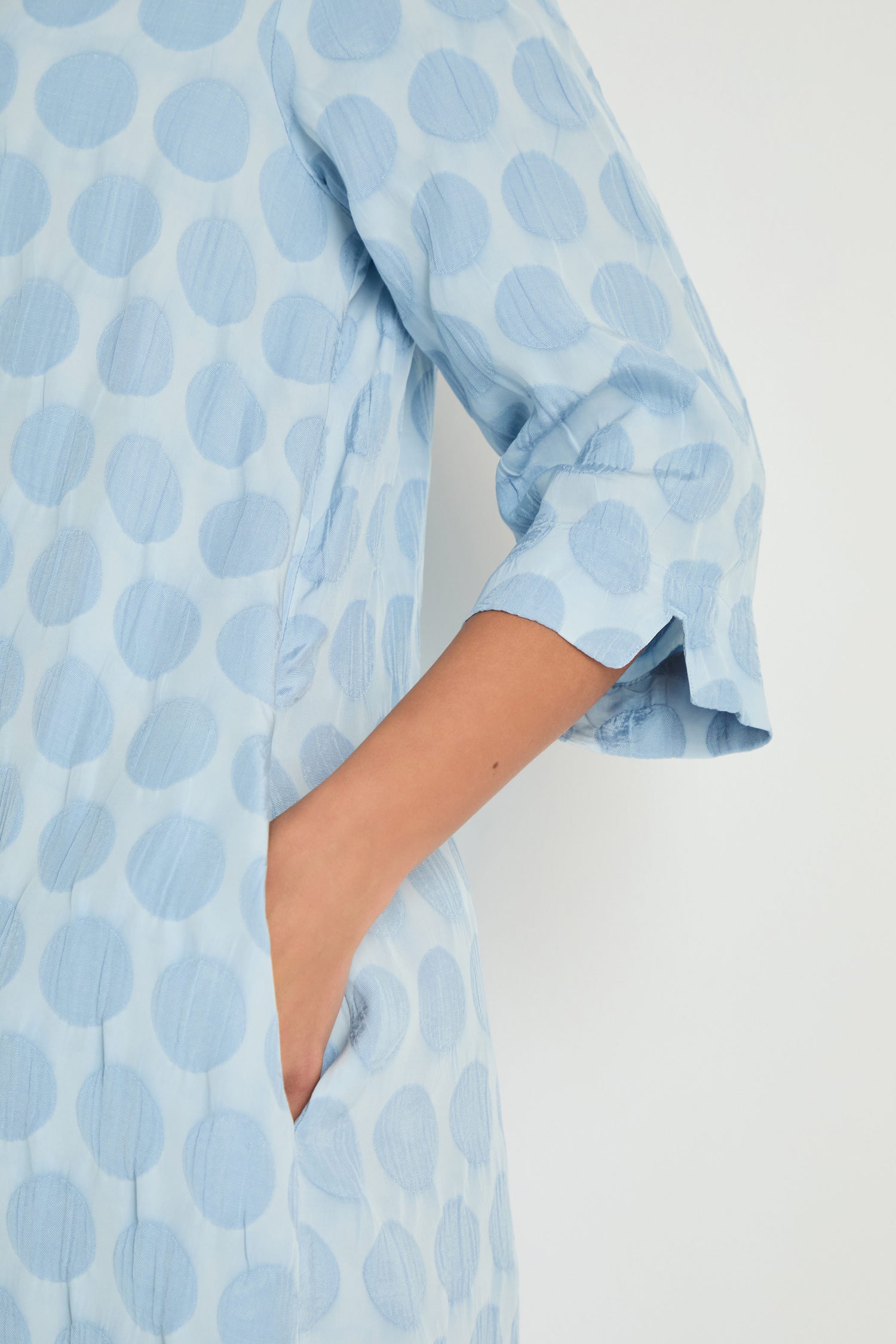 A person wears the Spot Textured Jacquard Bubble Dress in light blue, featuring long sleeves, large polka dots, midi length, and a side pocket where their right hand rests.