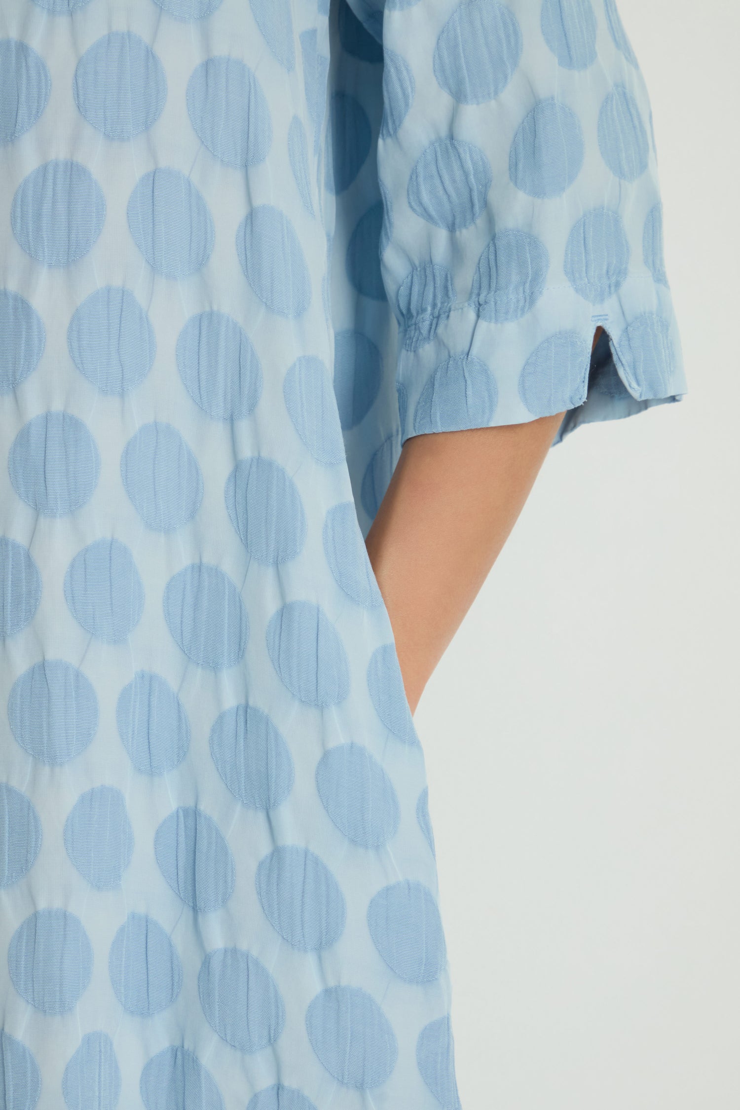 A person wears the Spot Textured Jacquard Bubble Dress in light blue, featuring long sleeves, large polka dots, midi length, and a side pocket where their right hand rests.