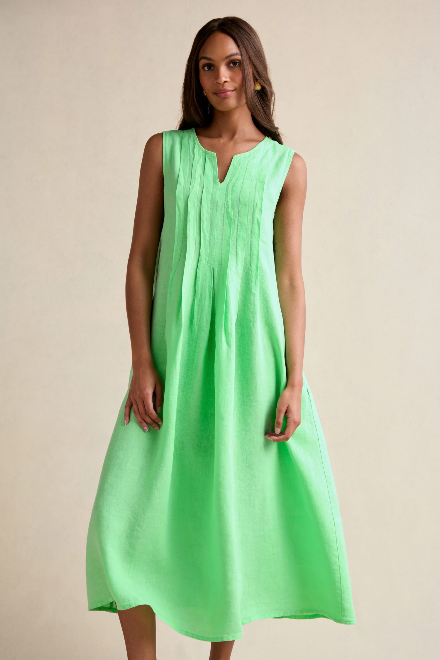 A woman faces forward, wearing the Linen Pleat Front Sleeveless Dress in mint green, a loose-fitting summer staple with pleat front details, set against a plain beige background.