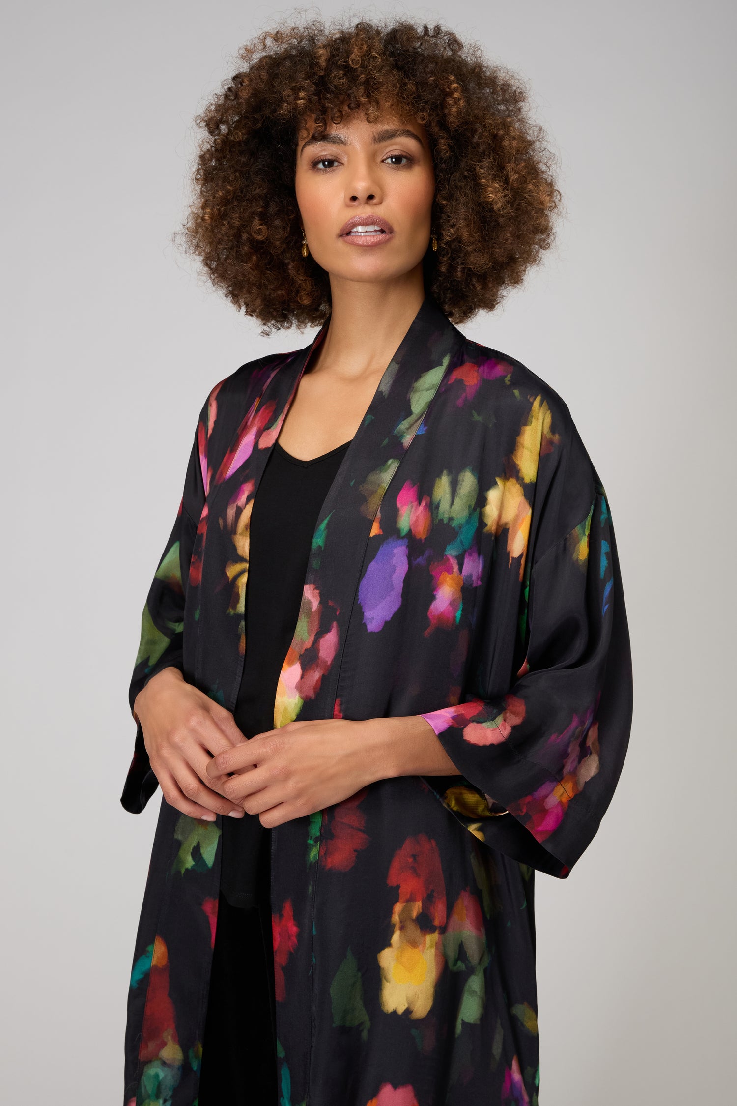 A woman with curly hair wears the Winter Floral Kimono, a black lightweight piece featuring a painterly floral print, over a black top, standing against a neutral background.
