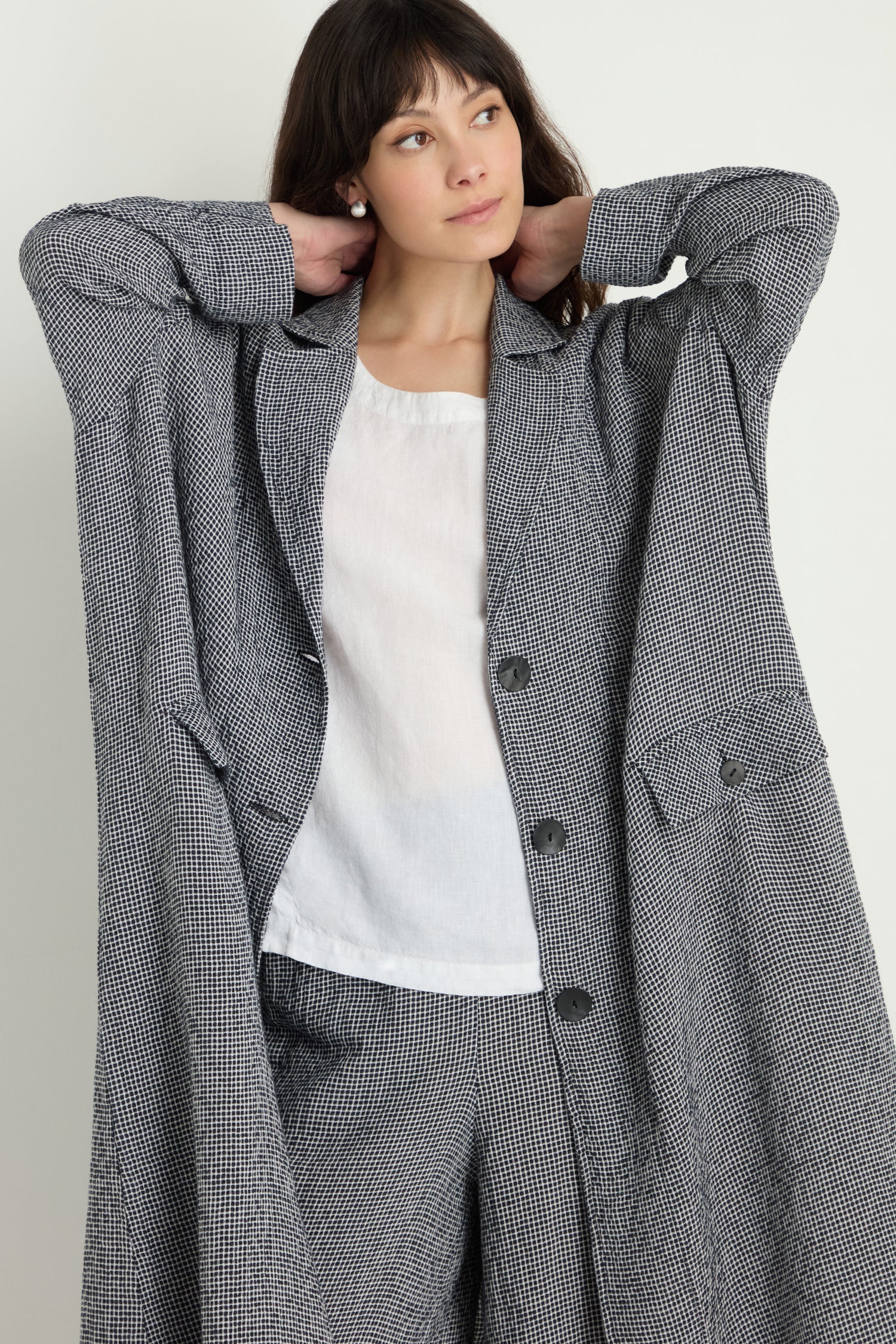 Woman wearing a long, oversized gray checked Micro Gingham Boxy Coat and matching pants over a white blouse, standing with her hands behind her head, looking slightly to the side—like fashion styled after electronics.
