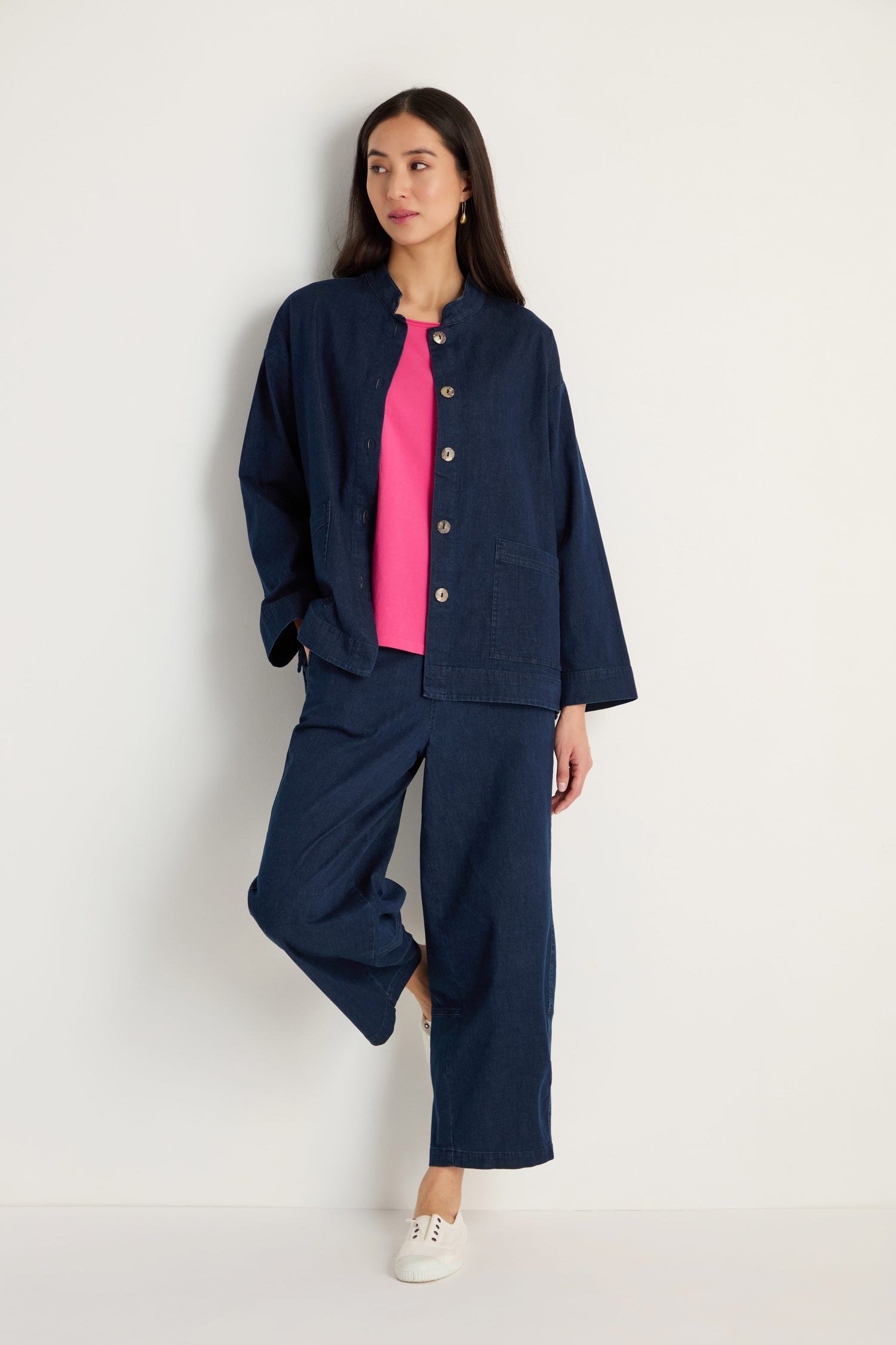 A woman leans against a white wall wearing the Stretch Denim Boxy Jacket and matching pants, paired with a pink top and white shoes.