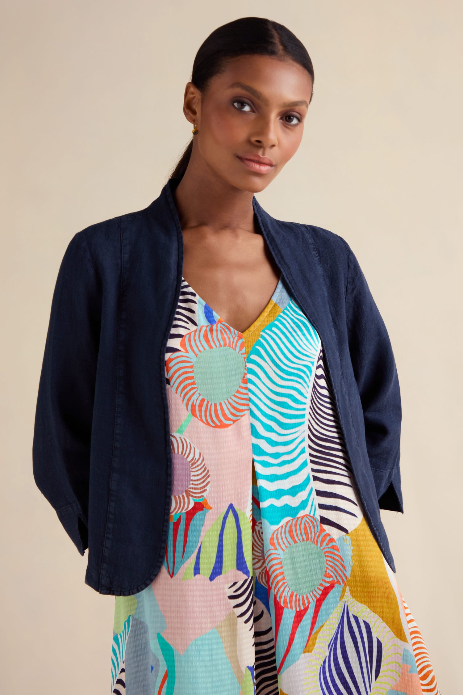 A woman with dark hair in a low ponytail wears the Navy Linen Cropped Jacket over a colorful, patterned dress, standing against a neutral background.