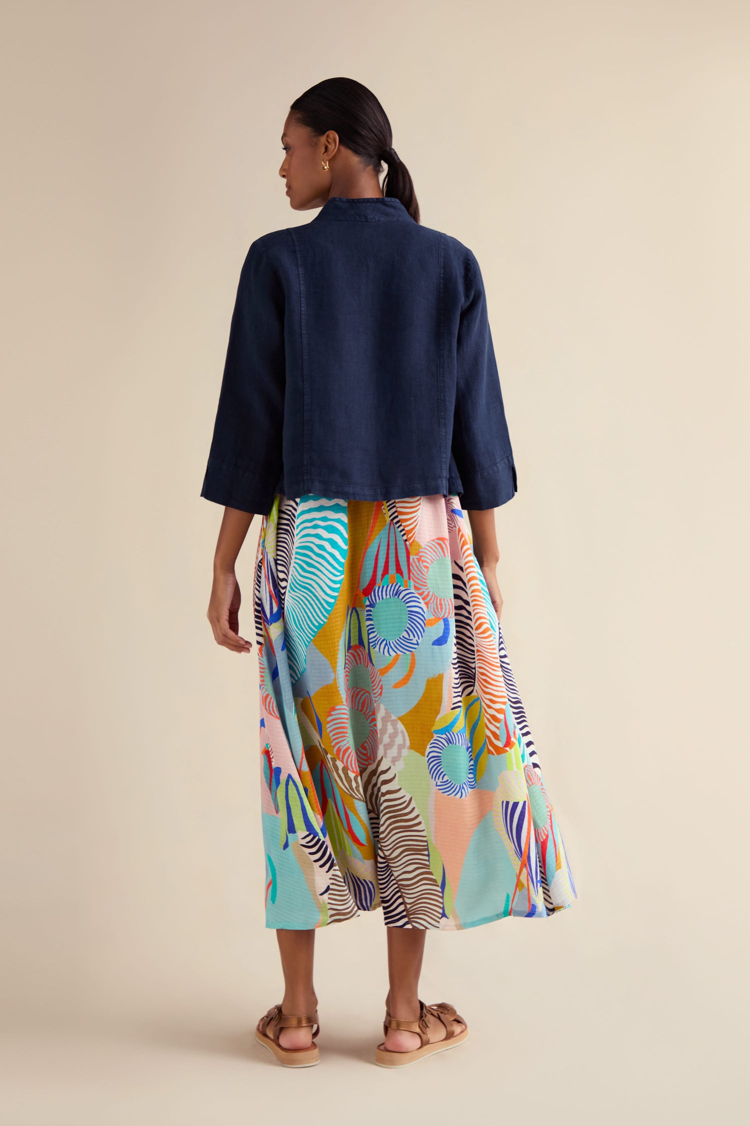 A woman faces away, wearing a boxy-fit Navy Linen Cropped Jacket with a colorful patterned skirt and tan sandals against a plain beige background.