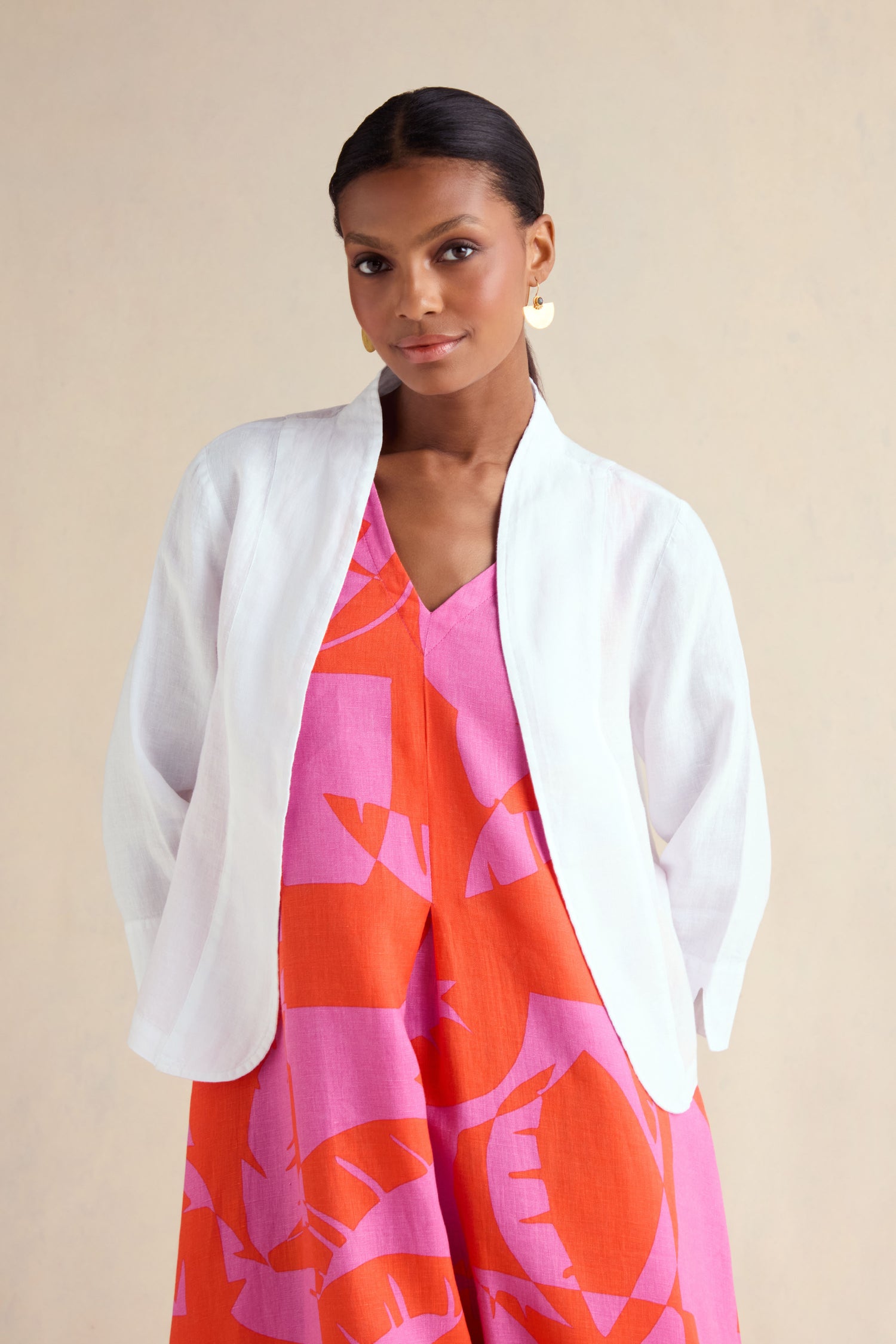 A woman stands against a plain light background, wearing the White Linen Cropped Jacket over a pink and orange patterned dress, showcasing this effortless layering piece.