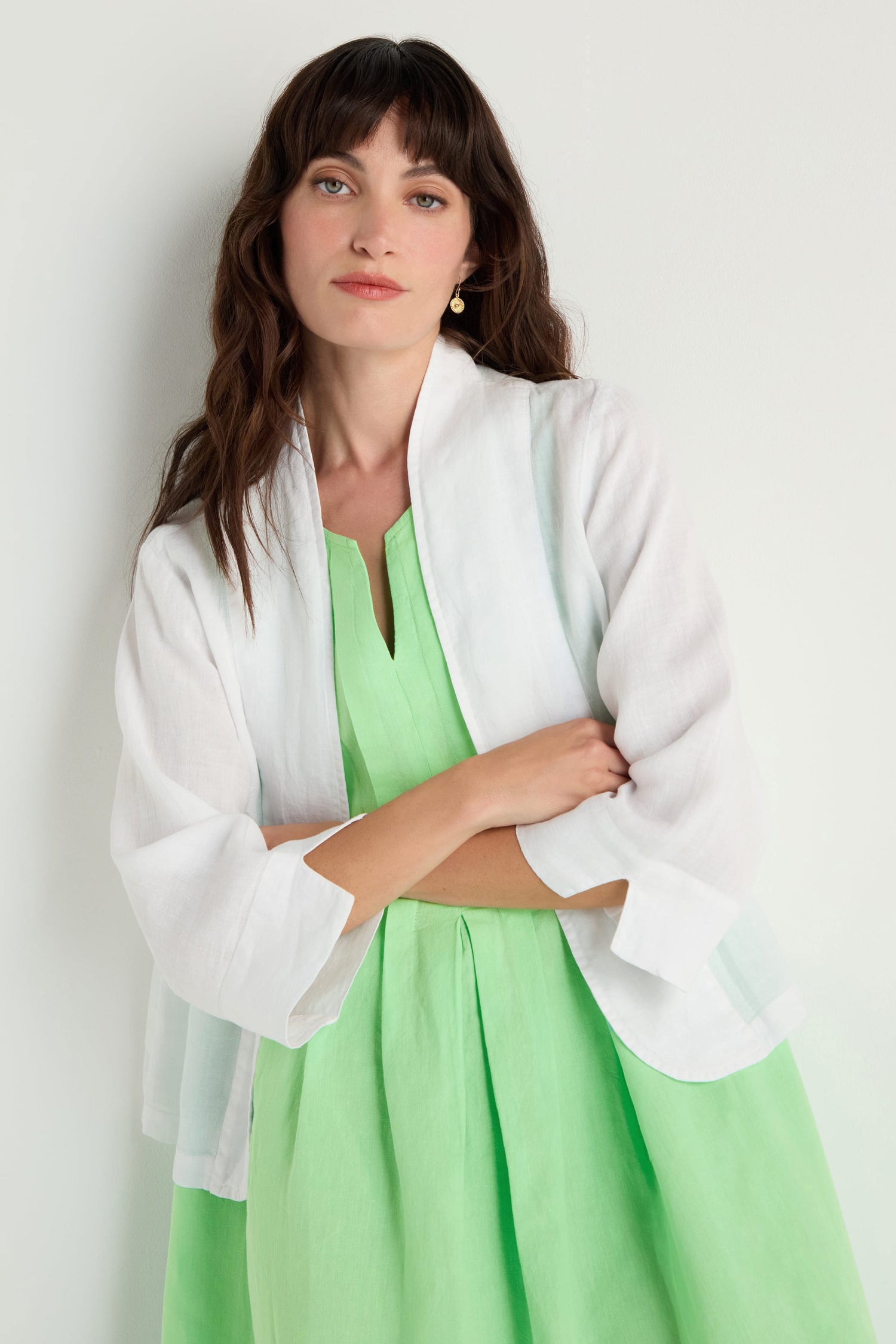 A woman with long brown hair wears a light green dress, a white sheer shawl, and the White Linen Cropped Jacket, featuring an edge-to-edge front and boxy fit, standing against a plain white background with her arms crossed.