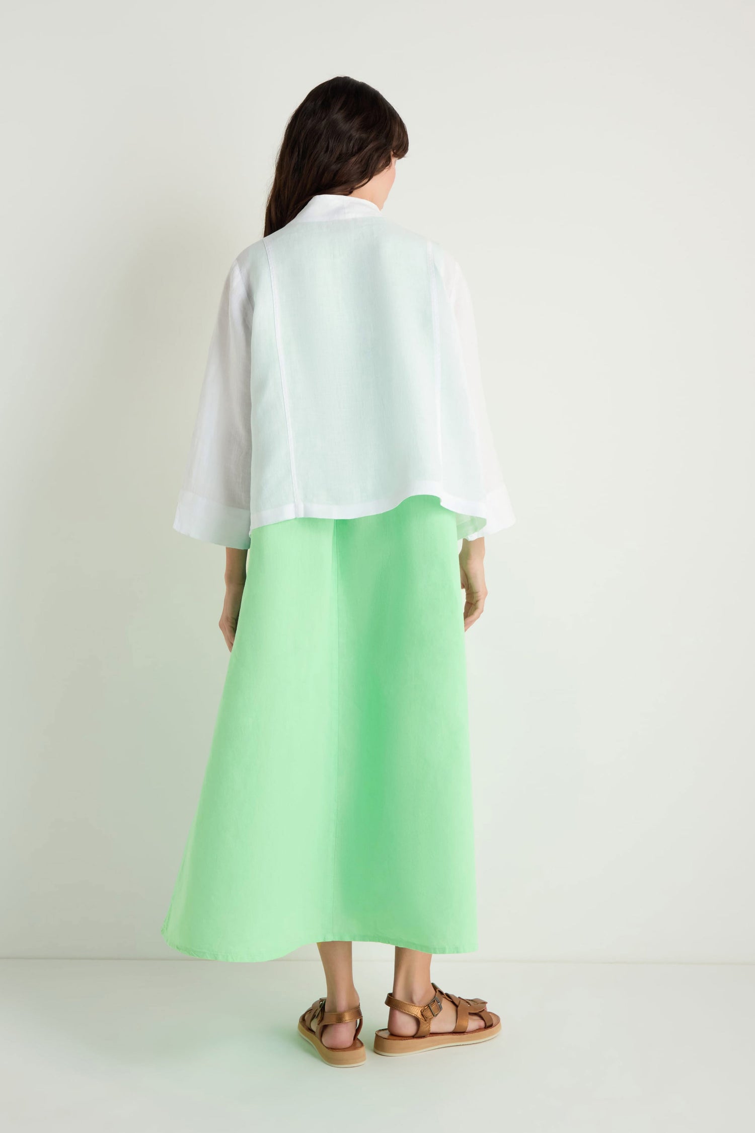 A person stands facing away, wearing a white linen cropped jacket over a white long-sleeve top, paired with a light green ankle-length skirt and brown sandals against a plain light background.
