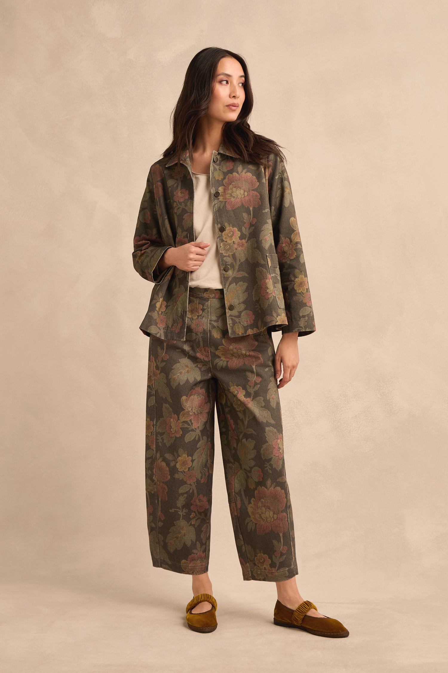 Woman wearing the Heirloom Damask Denim Jacket featuring a vintage archive print floral pattern in olive green, paired with matching wide-leg pants, a beige top, and mustard yellow shoes, standing against a beige background.