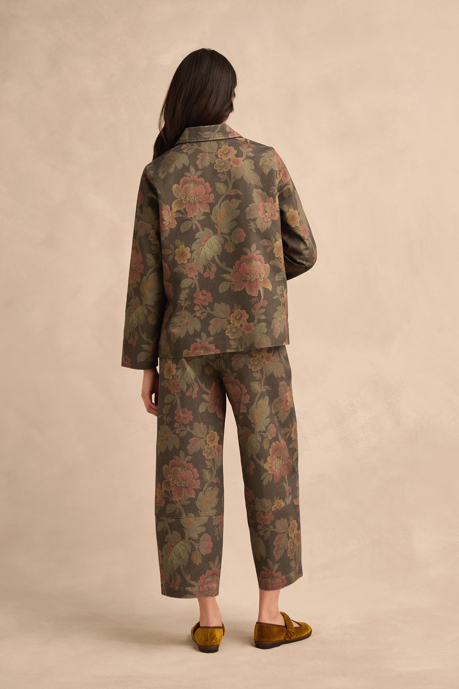 A person stands facing away, wearing the Heirloom Damask Denim Jacket with cropped pants and gold flats, against a neutral beige background.