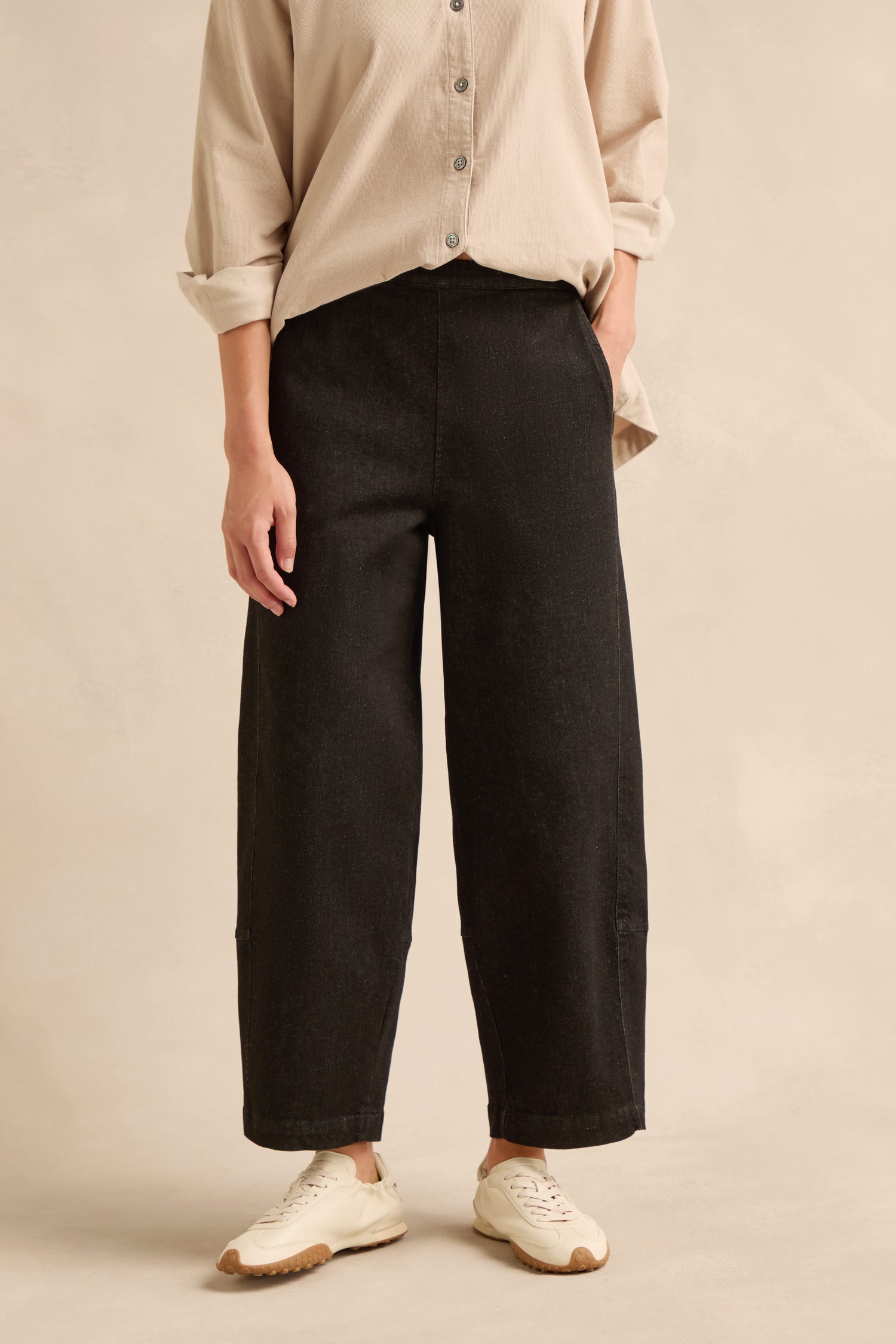 A person wearing a beige button-up shirt, Stretch Black Denim Bubble Trousers, and white sneakers stands against a neutral background.