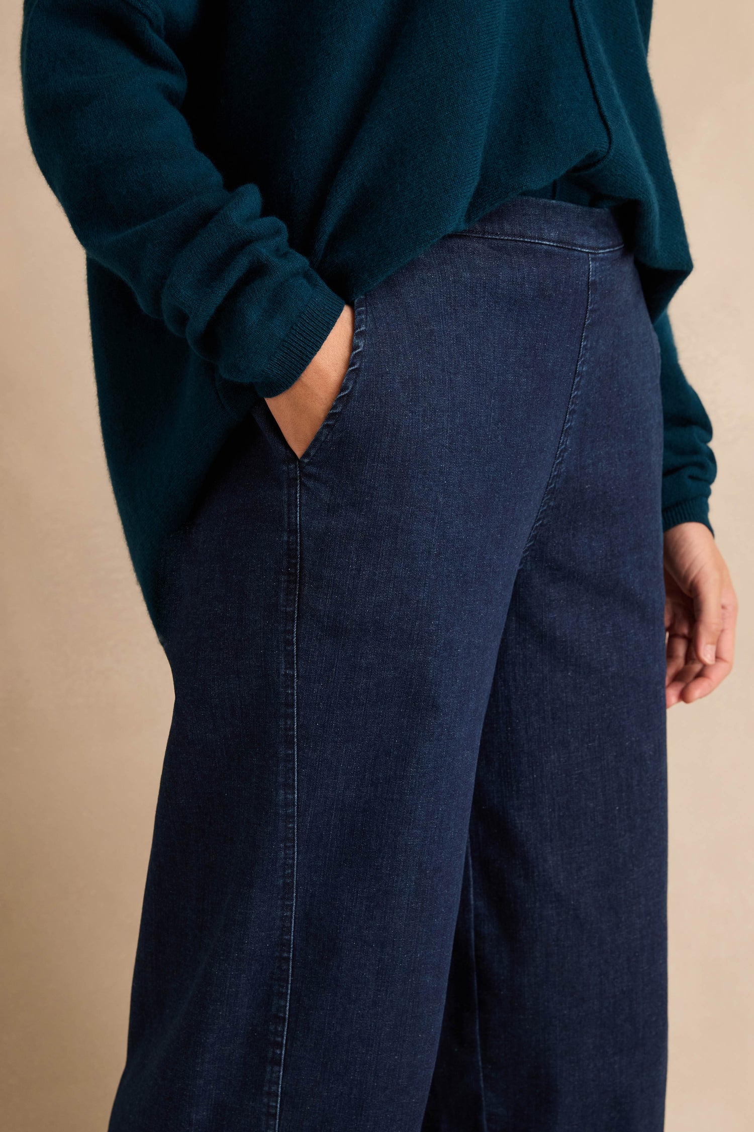 Wearing Stretch Denim Blue Bubble Trousers and a dark green sweater, the person stands with one hand in a pocket against a neutral background.