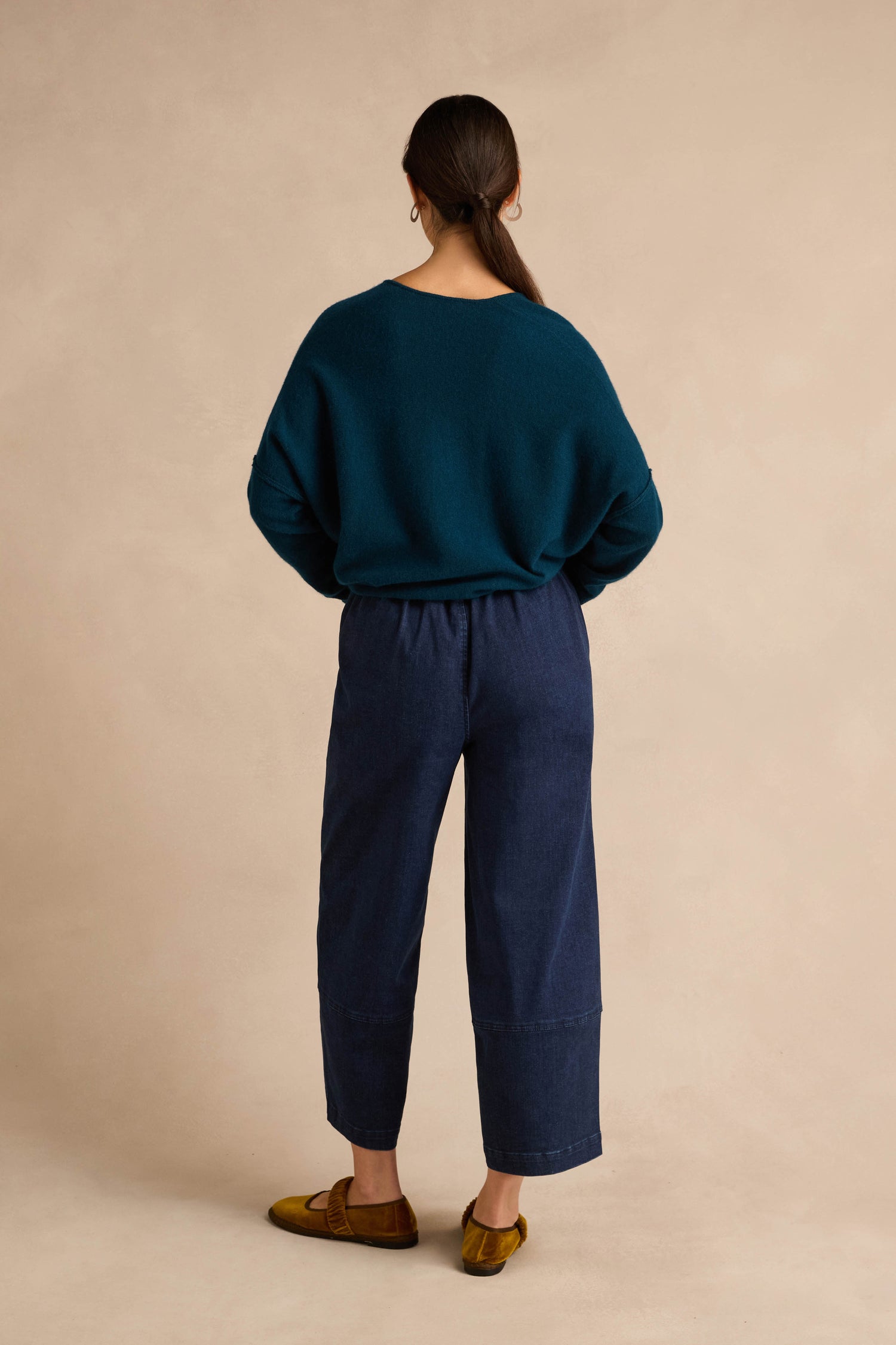 Person standing with back to camera, wearing a teal sweater, the Stretch Denim Blue Bubble Trousers, which are dark blue wide-leg pants with an elasticated waist, and brown shoes, against a beige background.