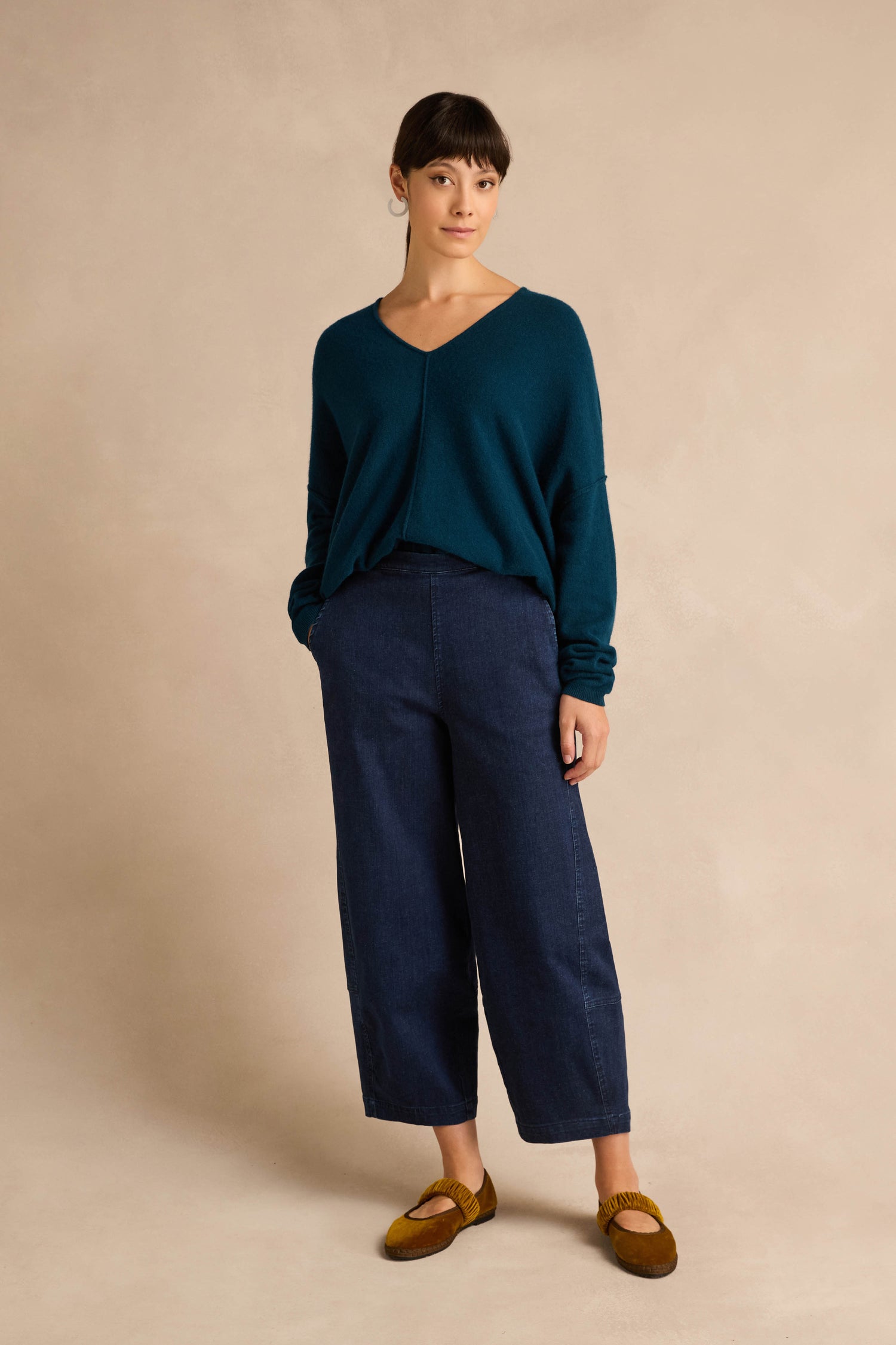 A woman stands against a beige backdrop wearing a teal V-neck sweater, Stretch Denim Blue Bubble Trousers, and brown shoes, with her hands in her pockets.