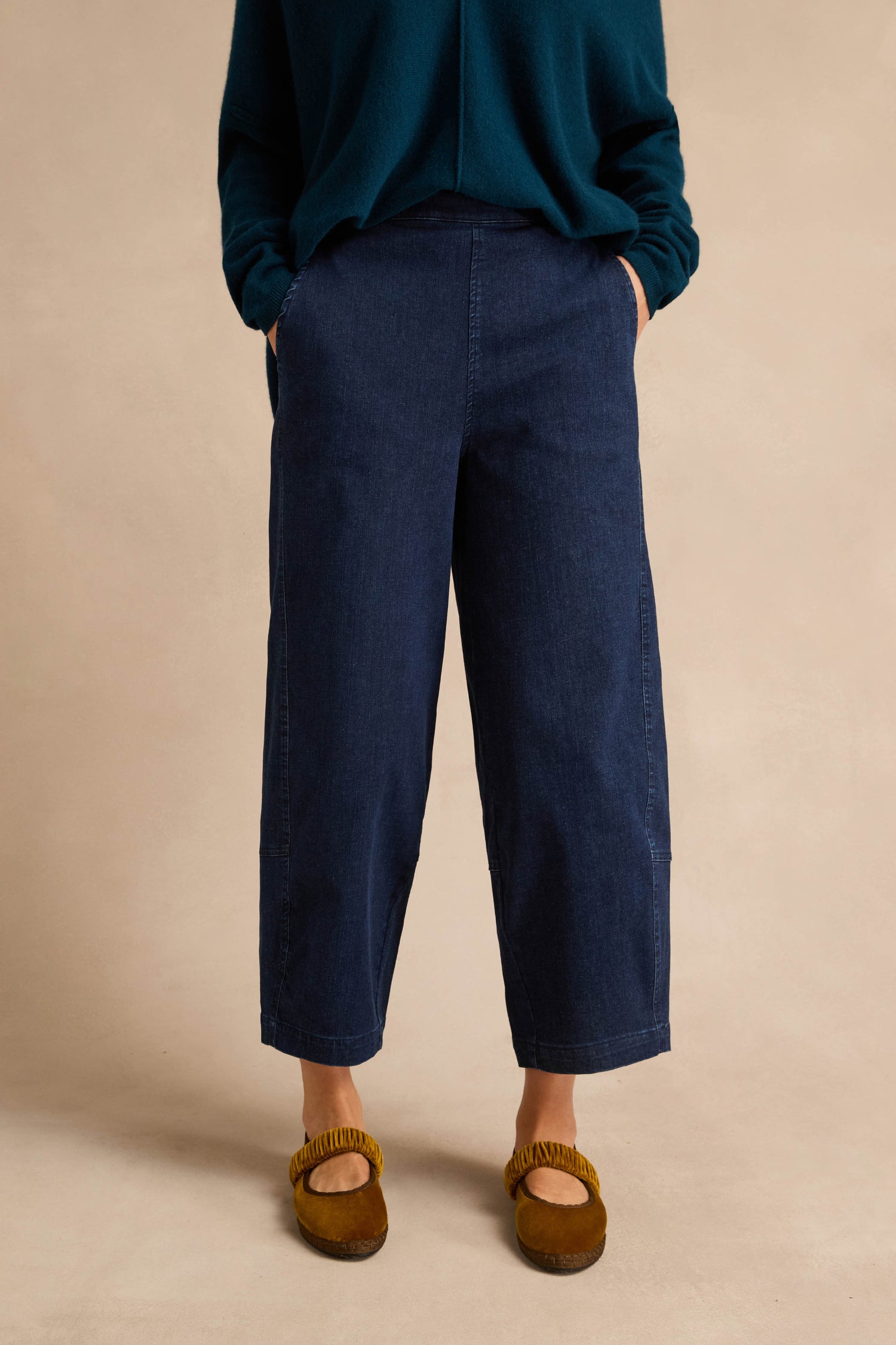 Wearing the Stretch Denim Blue Bubble Trousers—dark blue, wide-leg, cropped denim pants with an elasticated waist—paired with a dark green long-sleeve top and brown slip-on shoes, a person stands hands in pockets against a plain beige background.