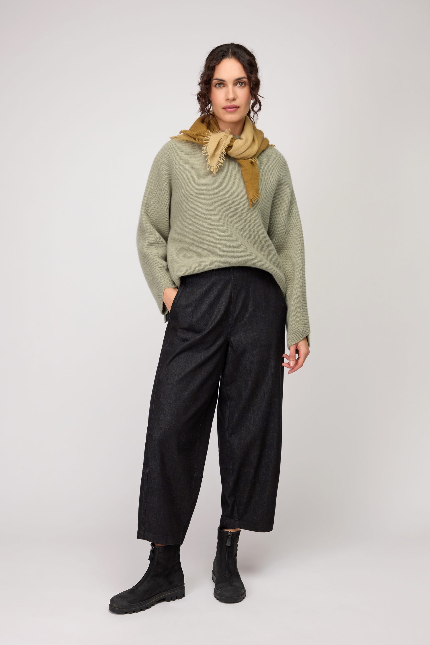 A woman stands against a plain background, dressed in a loose olive cocoon sweater, Black Stretch Denim Bubble Trousers, black boots, and a mustard scarf around her neck.