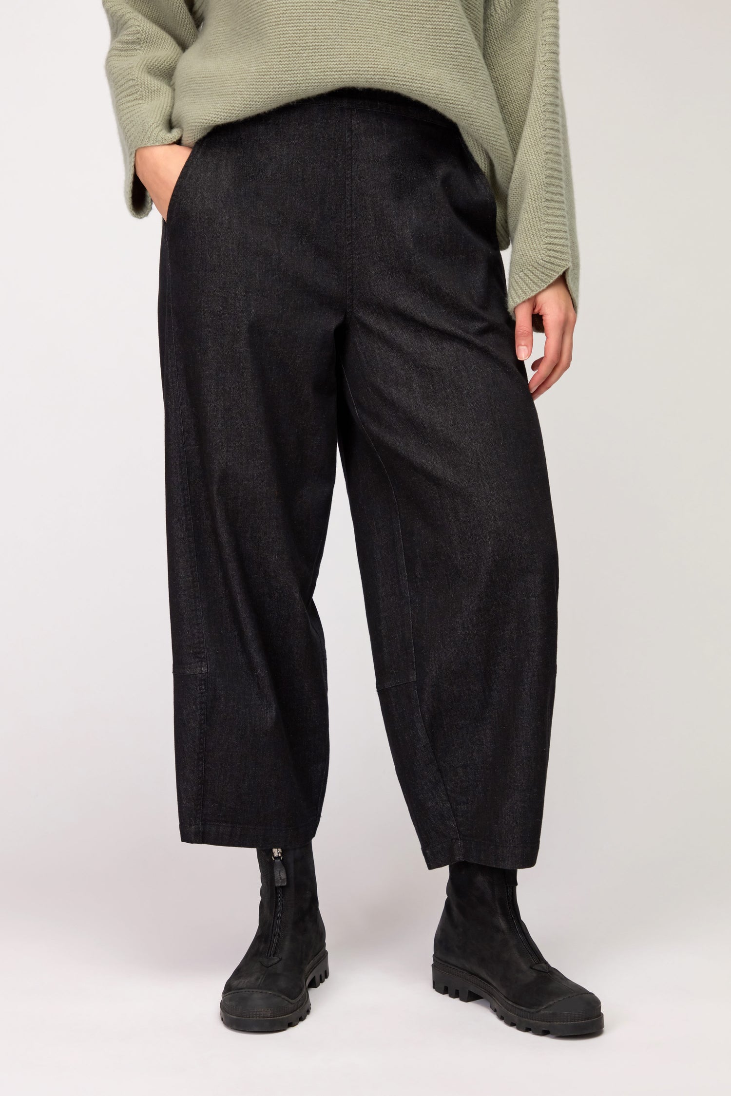 Person wearing Black Stretch Denim Bubble Trousers with a cocoon silhouette, paired with a loose light green sweater and black ankle boots, standing with one hand in a pocket against a plain background.