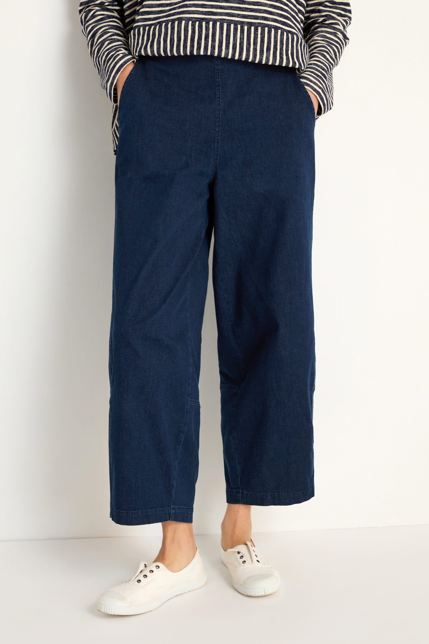 Person wears Stretch Denim Bubble Trouser in dark blue, paired with a striped long-sleeve shirt and white sneakers, standing against a plain background with hands in pockets.