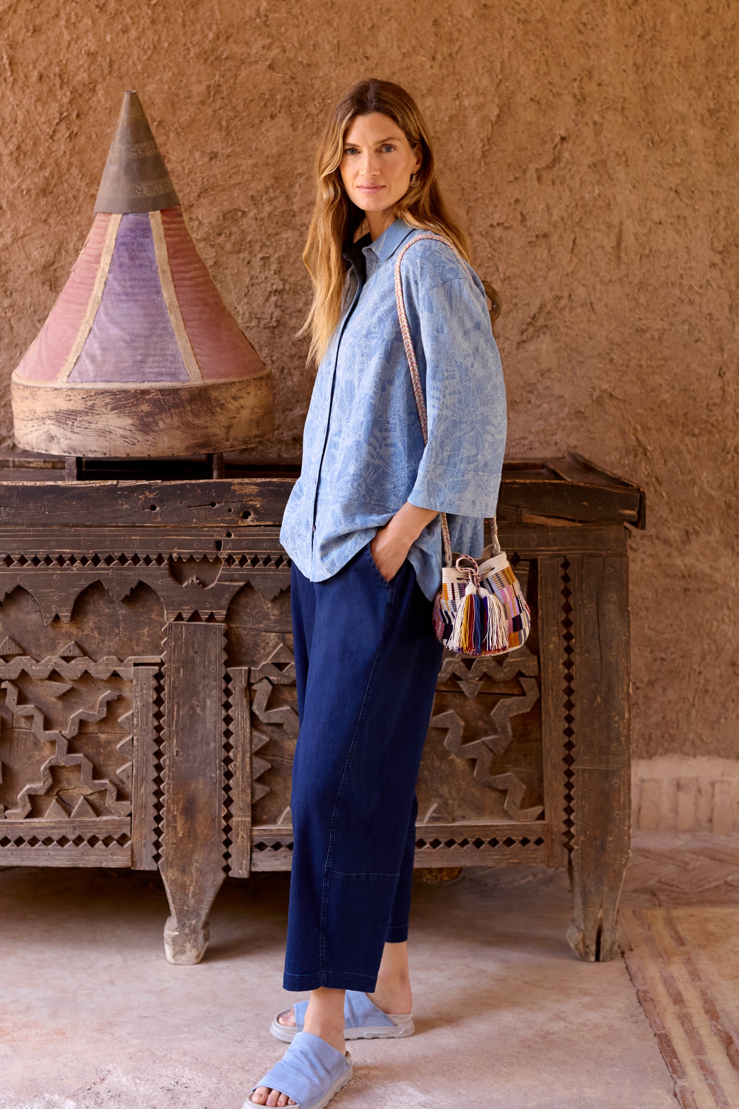 A woman stands indoors by a rustic table and textured wall, wearing a blue shirt, blue sandals, carrying a colorful tasseled bag, and featuring the Stretch Denim Bubble Trouser in a relaxed fit.