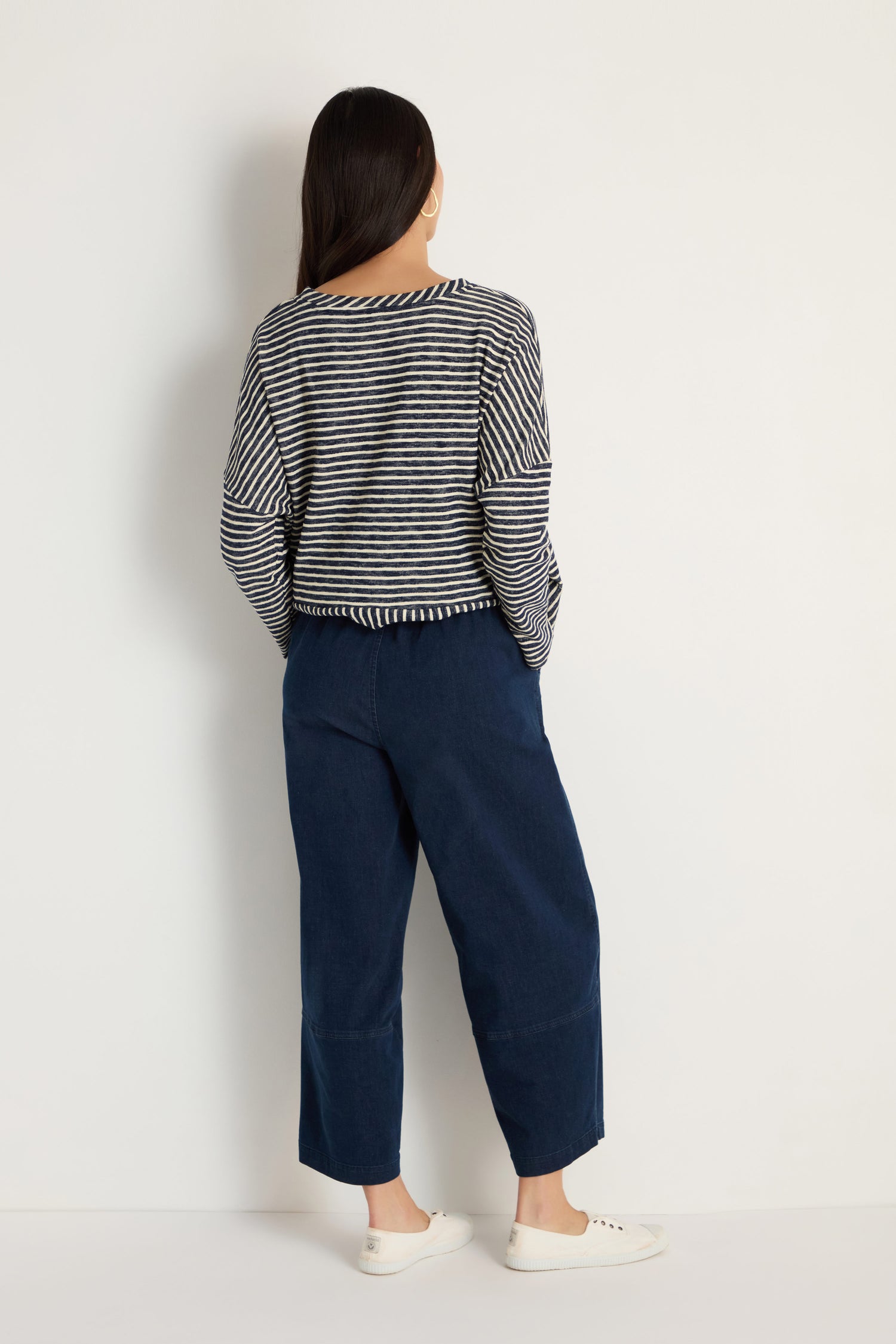 A person with long dark hair, wearing a black and white striped long-sleeve shirt, Stretch Denim Bubble Trousers in navy, and white sneakers stands facing a white wall with their back to the camera.