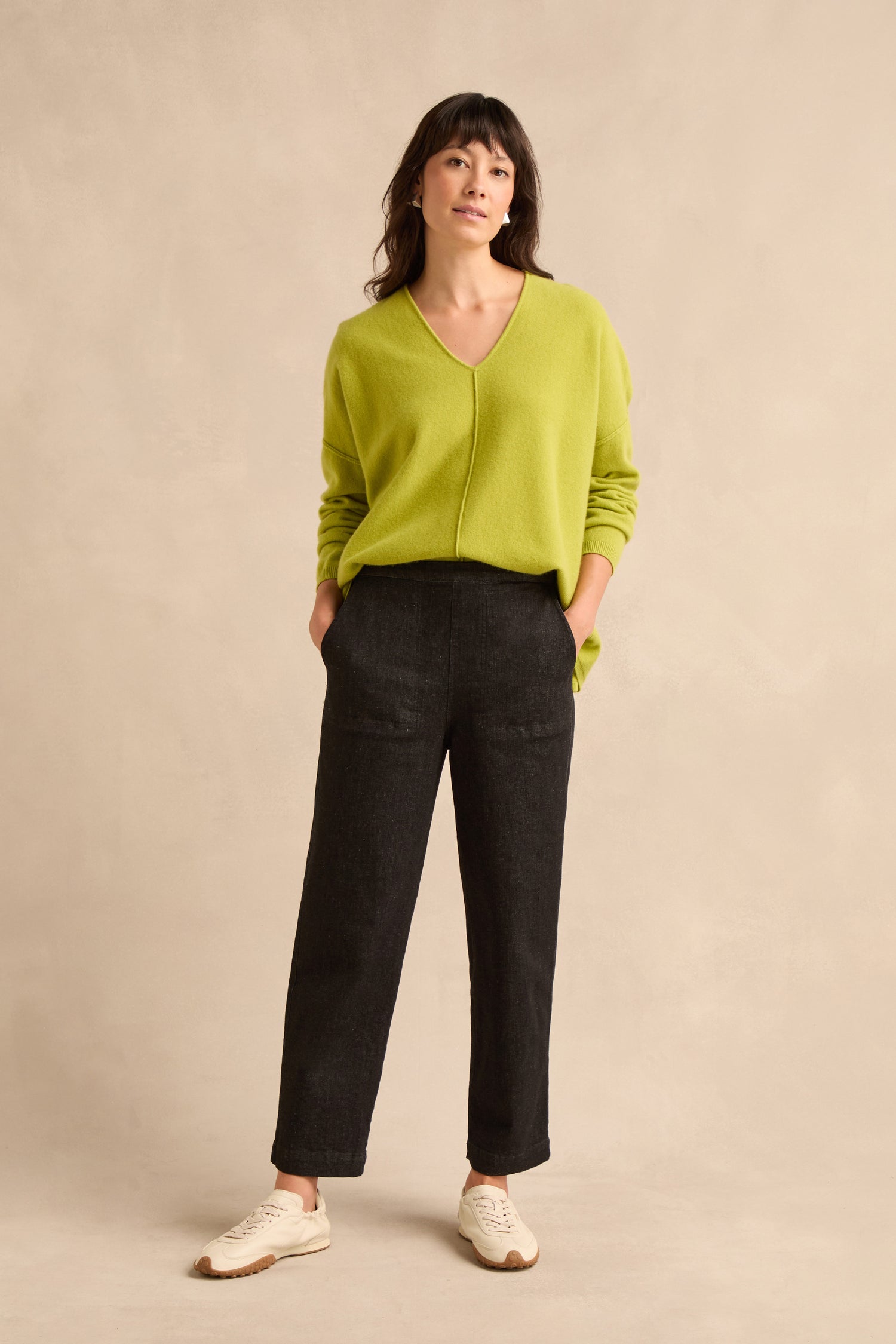 Woman standing with hands in pockets, wearing a lime green sweater, Stretch Black Denim Straight trousers with an elasticated waist, and white sneakers, against a beige background.