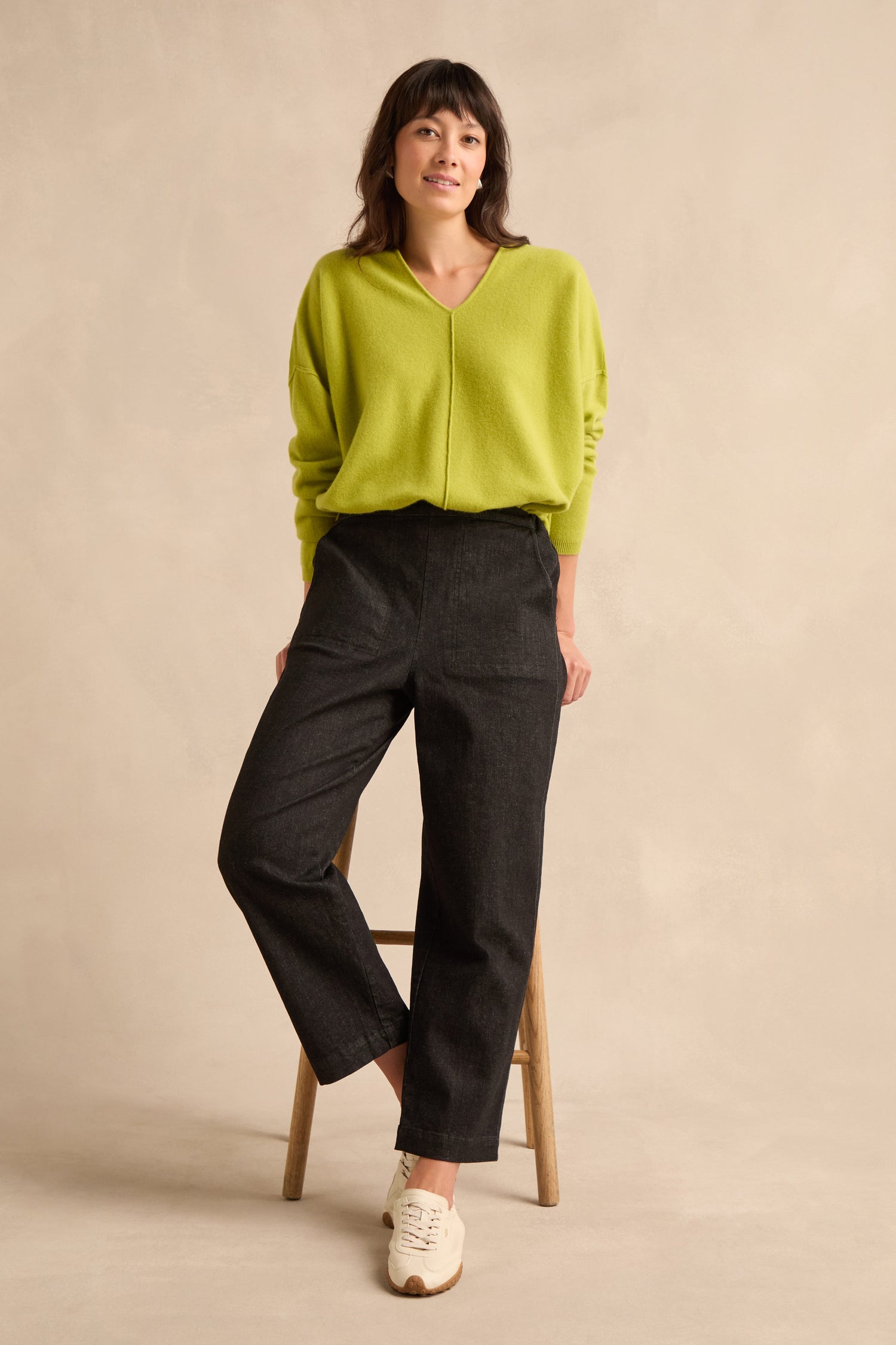 A person wearing a lime green sweater, Stretch Black Denim Straight trousers with an elasticated waist, and white sneakers stands with one leg on a wooden stool against a beige background.