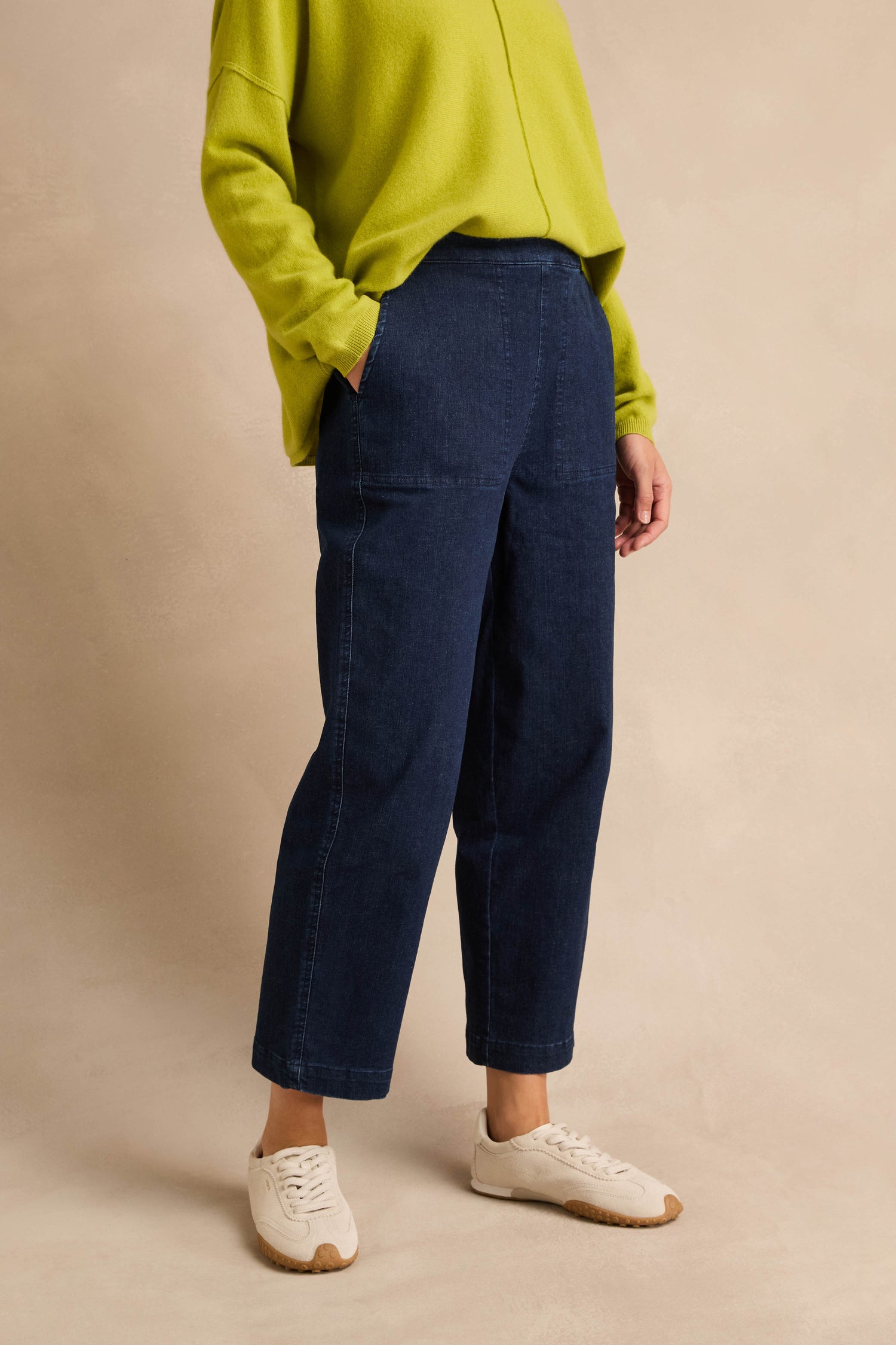 Person wearing a loose lime green sweater, Stretch Denim Blue Straight Trousers, and white sneakers, standing against a beige background.