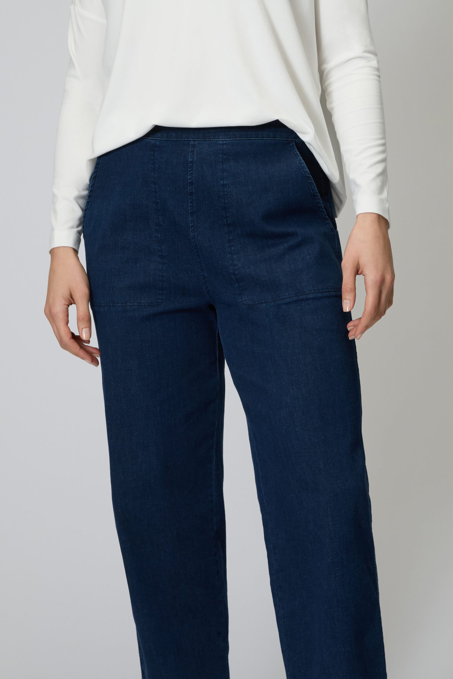 Model wears Stretch Denim Straight Trouser in dark blue with hands in pockets, paired with a long-sleeved white top. Shown from shoulders to ankles against a plain background.
