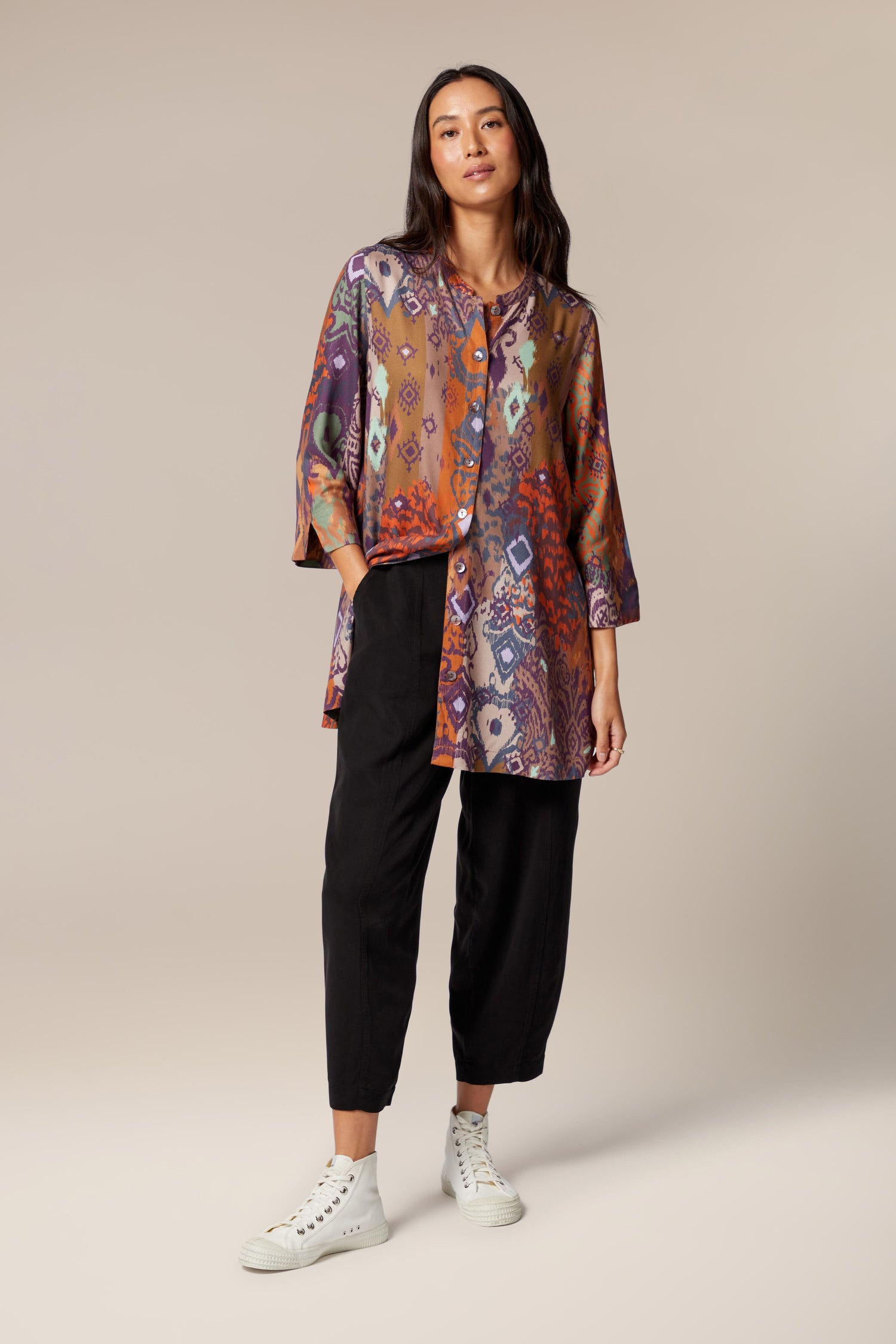 A model wears a printed button-up shirt, wide black trousers with tapered ankles. She wears neutral trainers.
