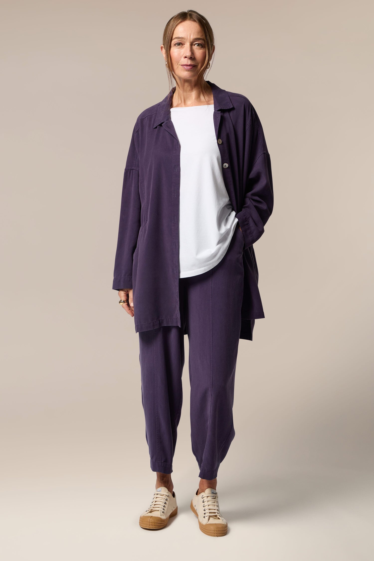 A model wears an open purple button-up shirt, white top underneath and purple wide trousers with tapered ankles. She wears neutral trainers.