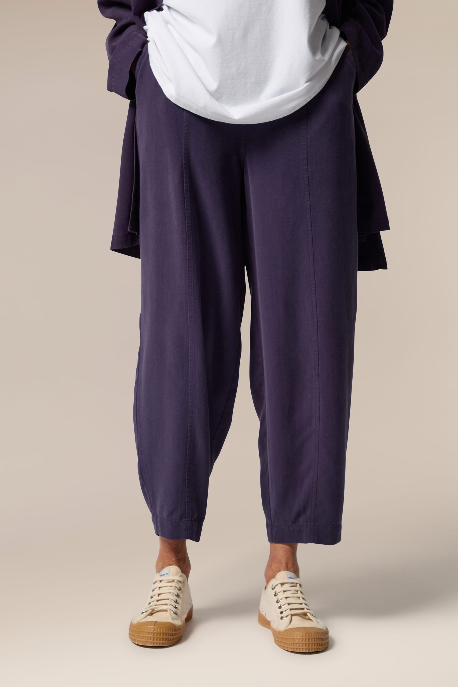 A model wears an open purple button-up shirt, white top underneath and purple wide trousers with tapered ankles. She wears neutral trainers.
