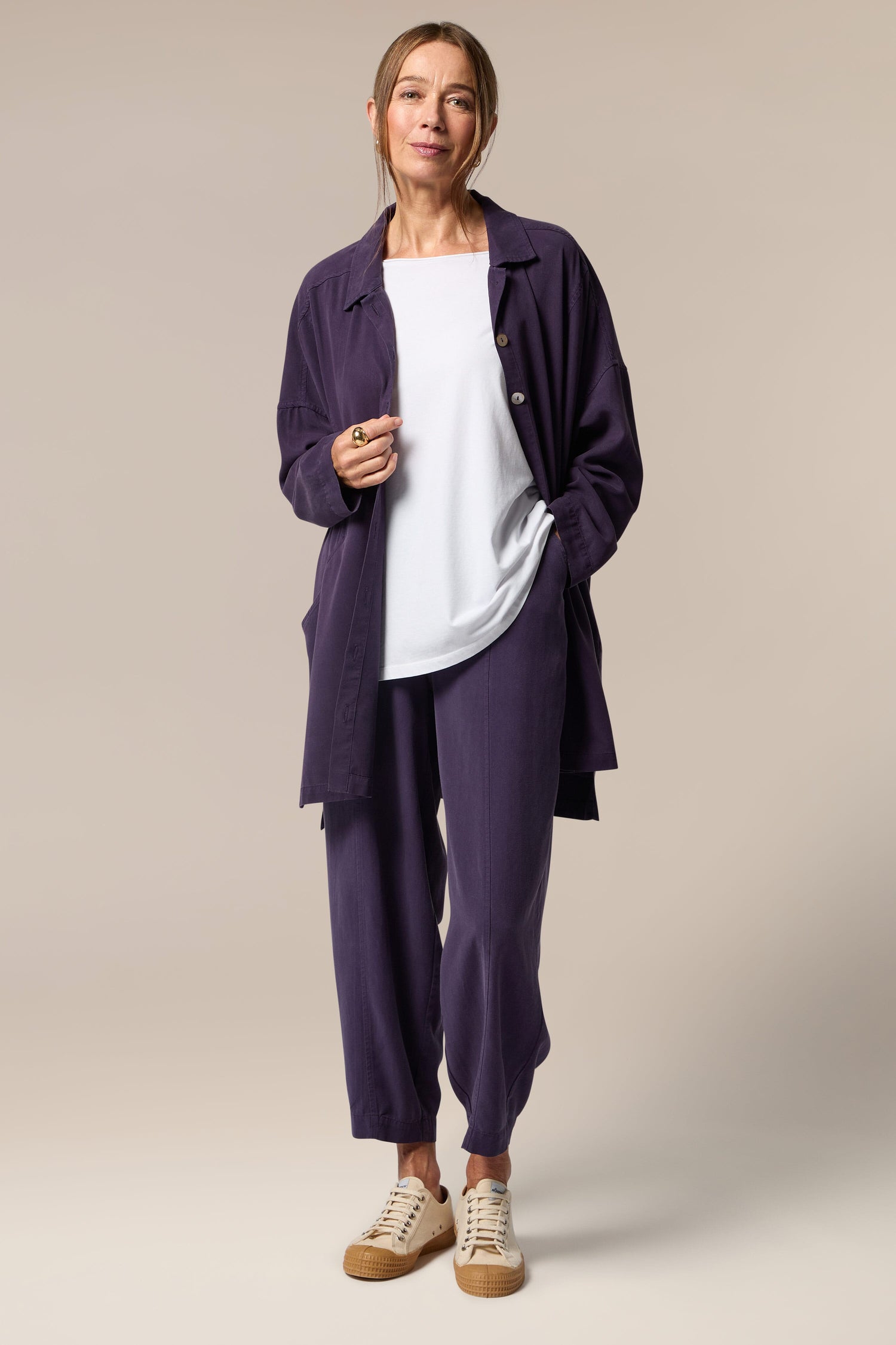 A model wears an open purple button-up shirt, white top underneath and purple wide trousers with tapered ankles. She wears neutral trainers.
