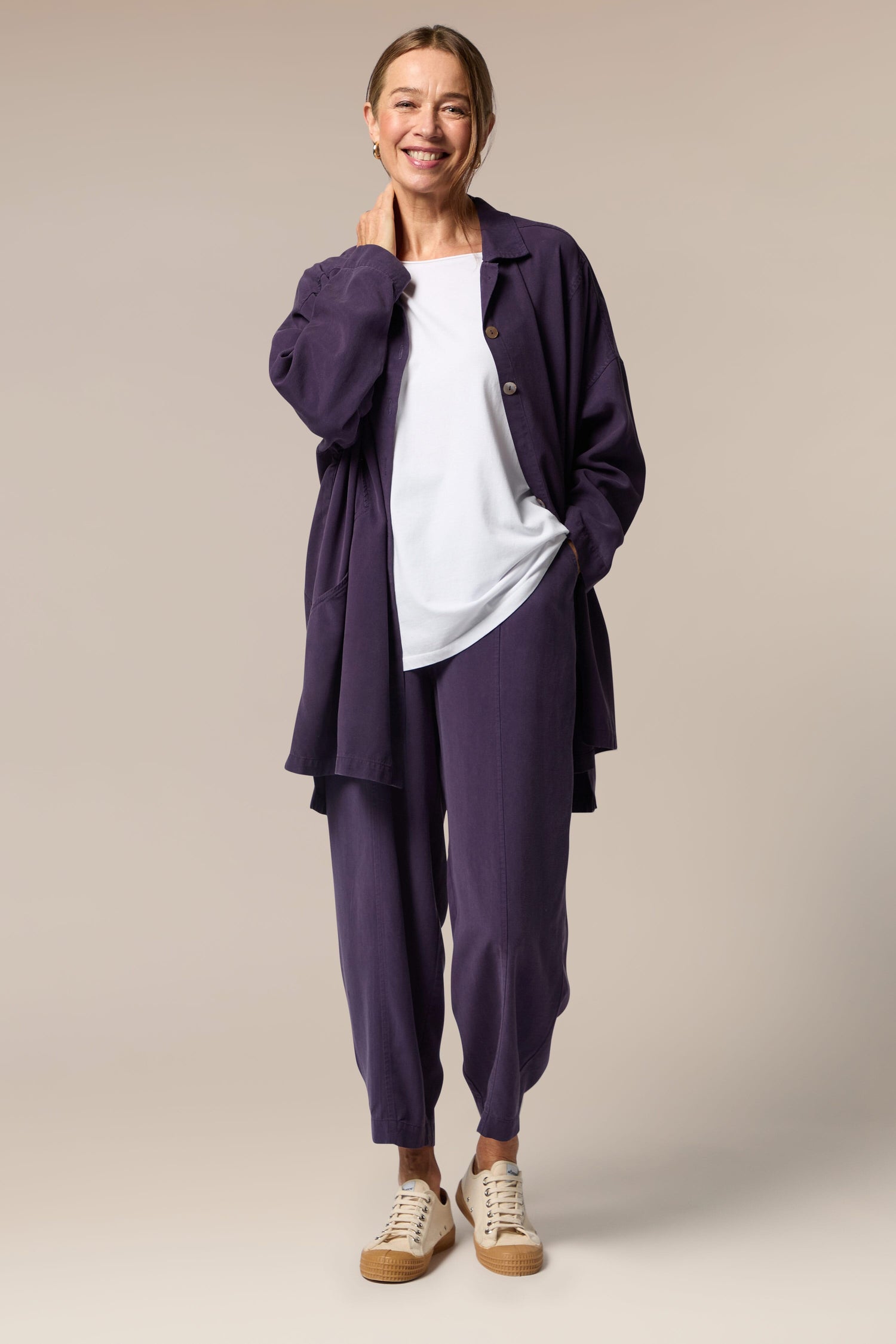 A model wears an open purple button-up shirt, white top underneath and purple wide trousers with tapered ankles. She wears neutral trainers.