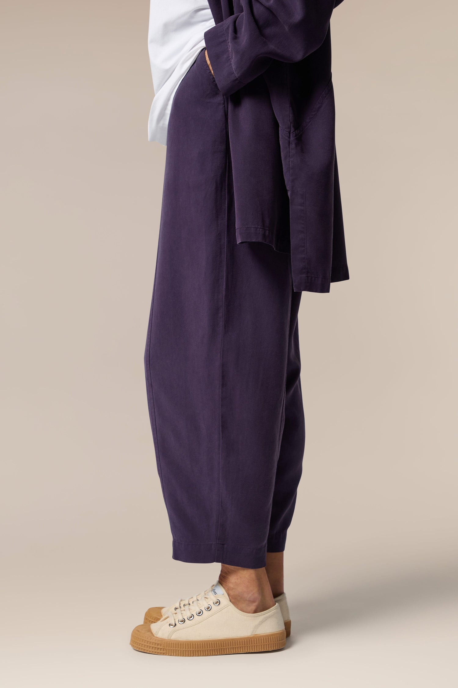 A model wears an open purple button-up shirt, white top underneath and purple wide trousers with tapered ankles. She wears neutral trainers.