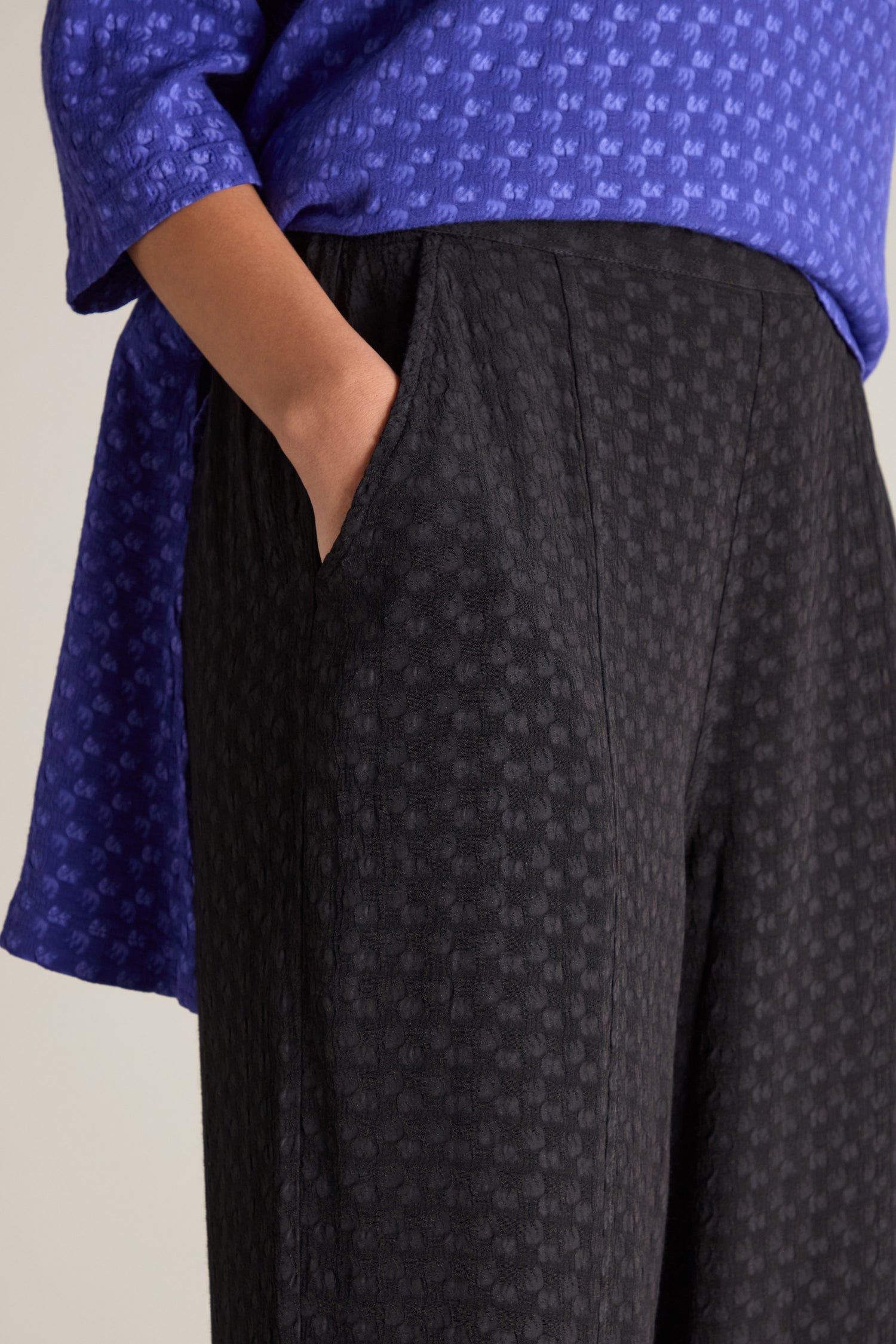 Person wearing a purple patterned top and textured black Small Square Jacquard Bubble Trousers, which feature an elasticated waistband, with one hand in the pocket.